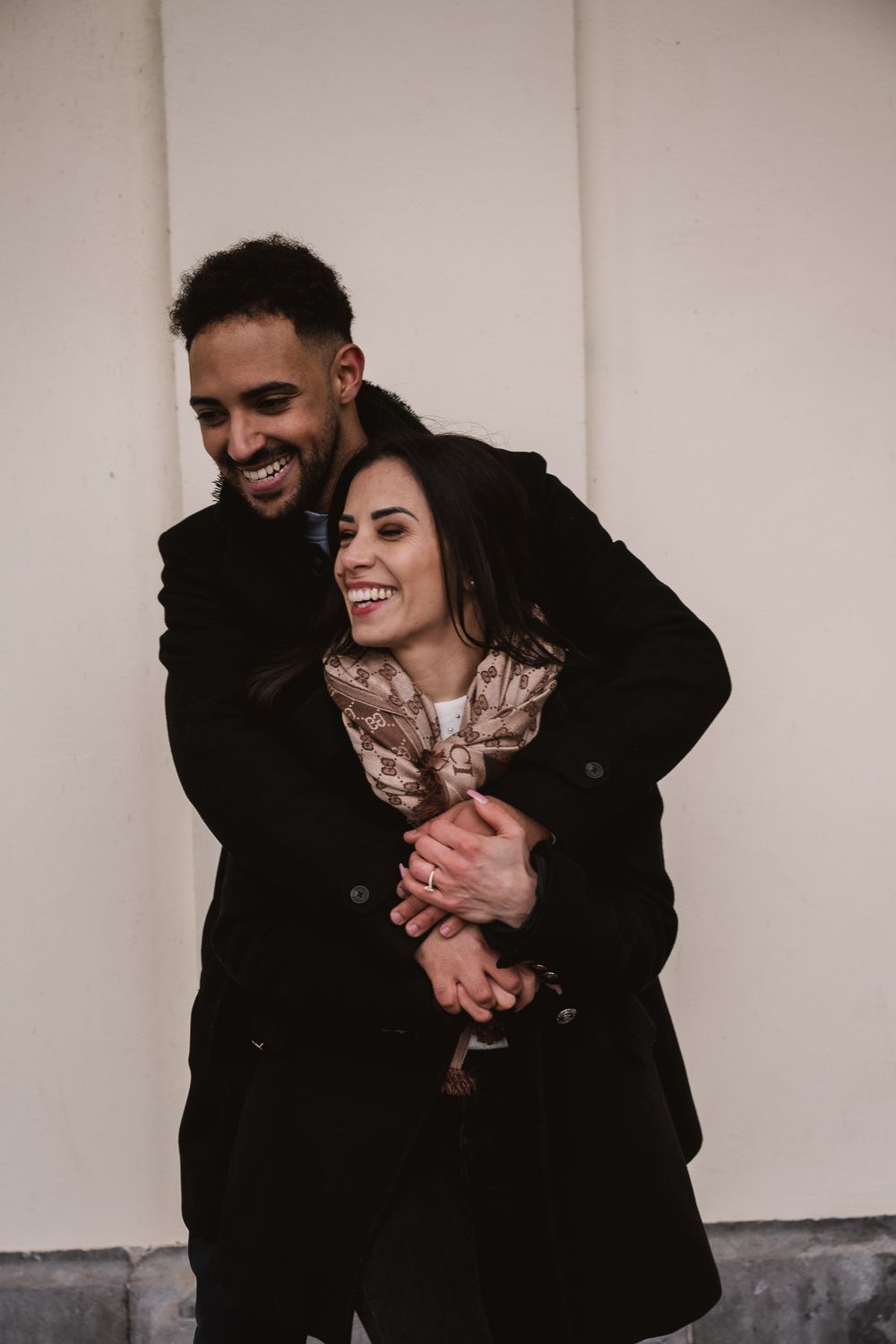 A romantic candid moment shows a couple embracing and laughing together against a light colored wall.