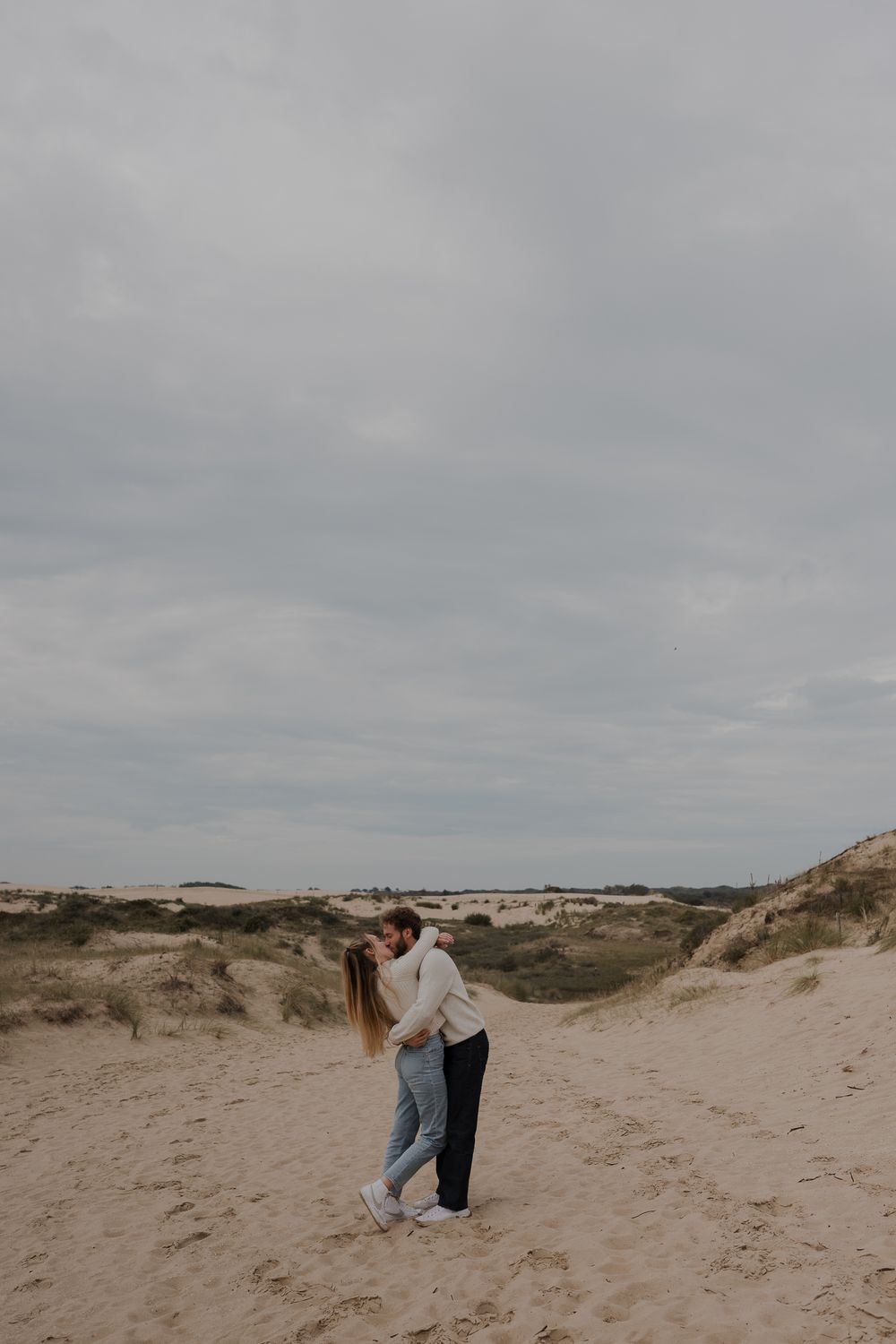 A couple embraces on a cloudy day among rolling sand dunes at the beach.