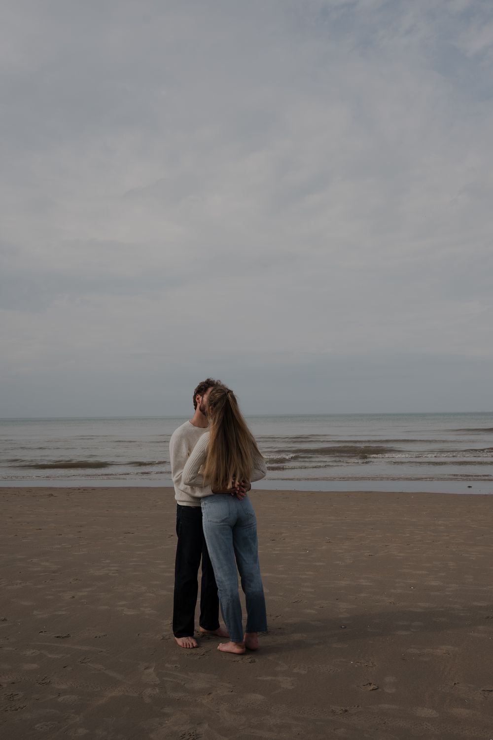 A couple embraces while standing barefoot on an empty beach under overcast skies.