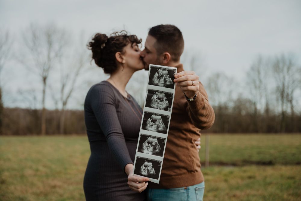A couple kisses while holding ultrasound photos in a field during autumn, announcing their pregnancy.