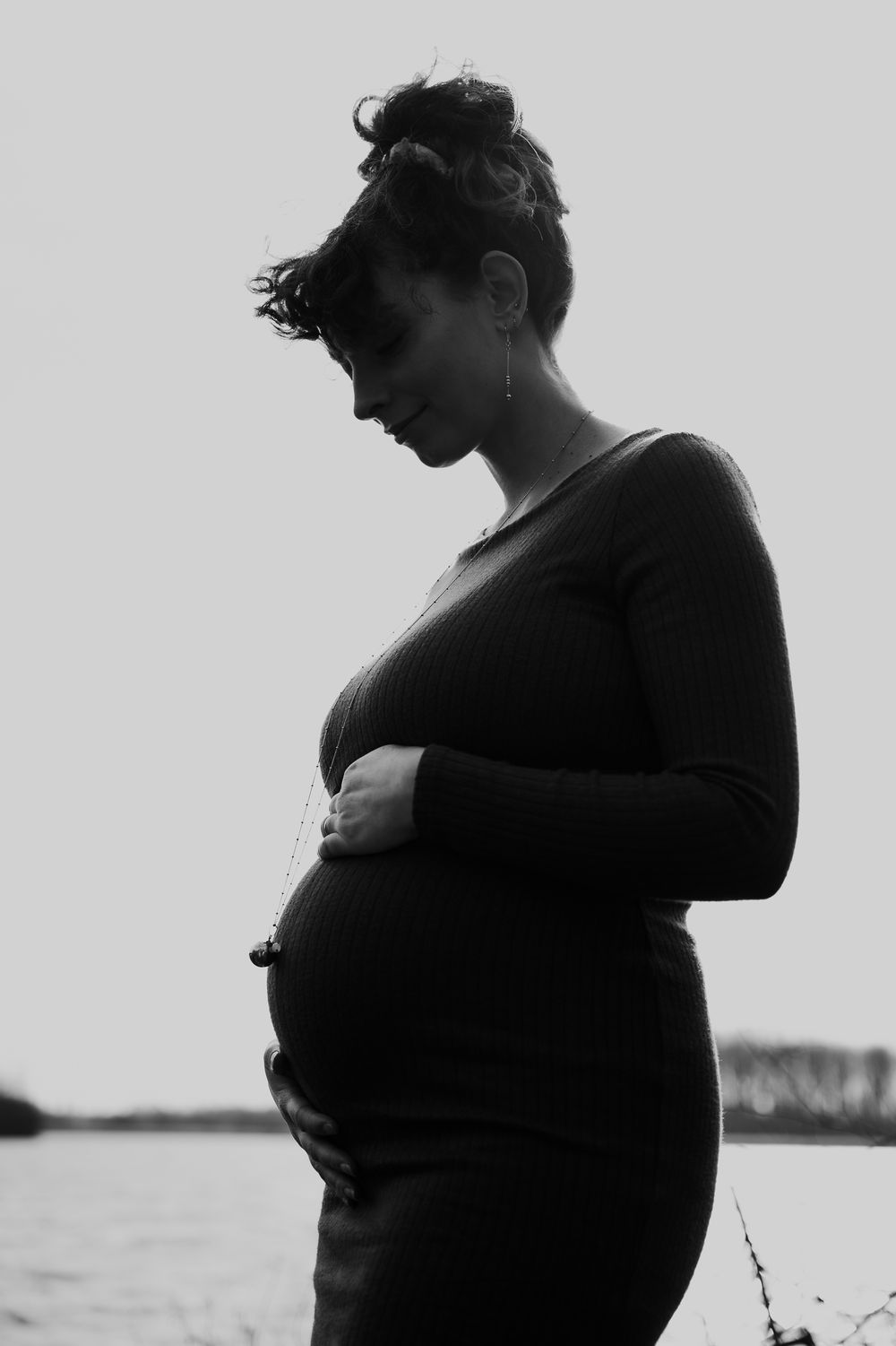 Black and white silhouette of a pregnant woman in profile standing by water, wearing a long sleeve dress.