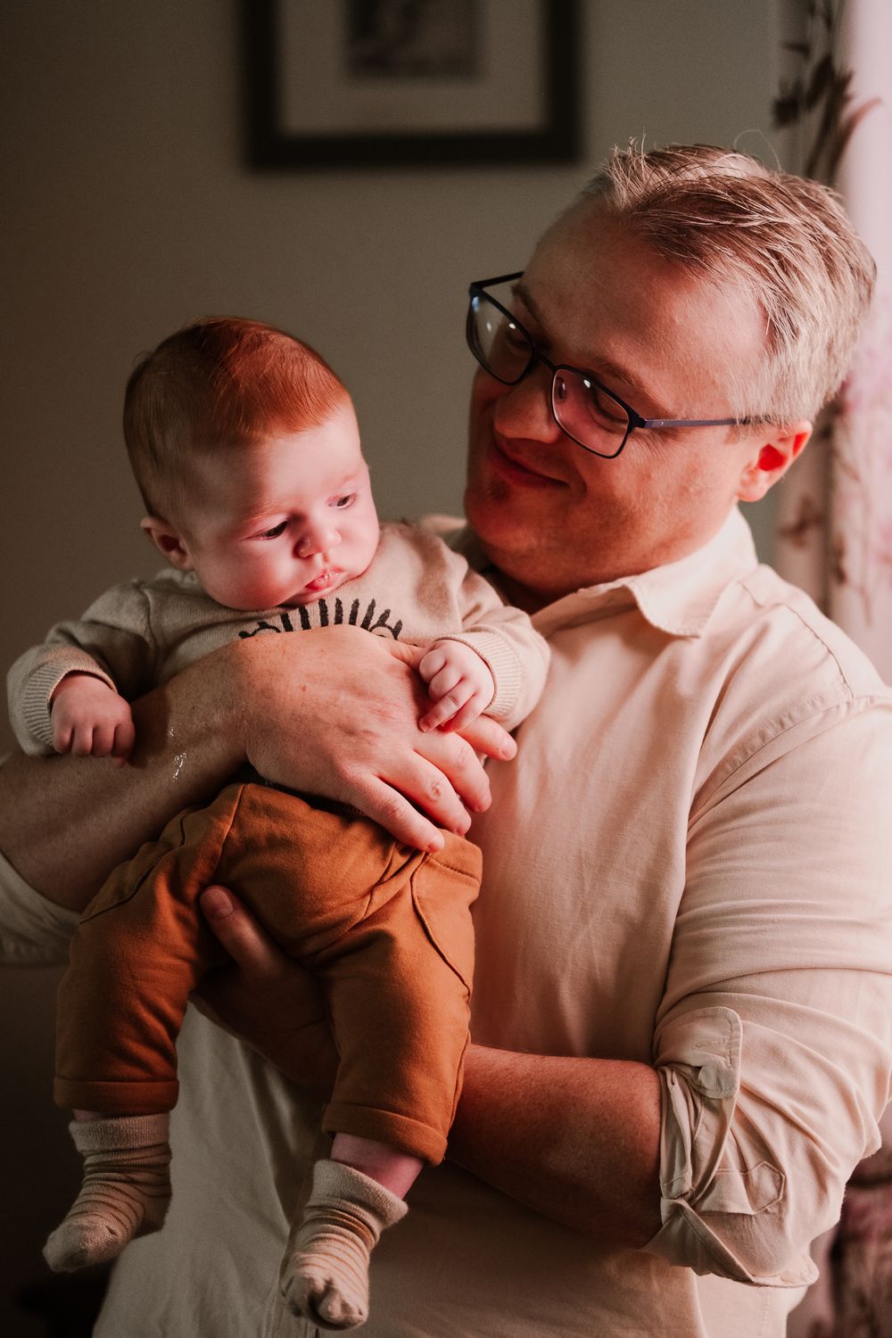 A tender moment captured between an adult in glasses wearing a white shirt and a baby dressed in brown pants and cream sweater.