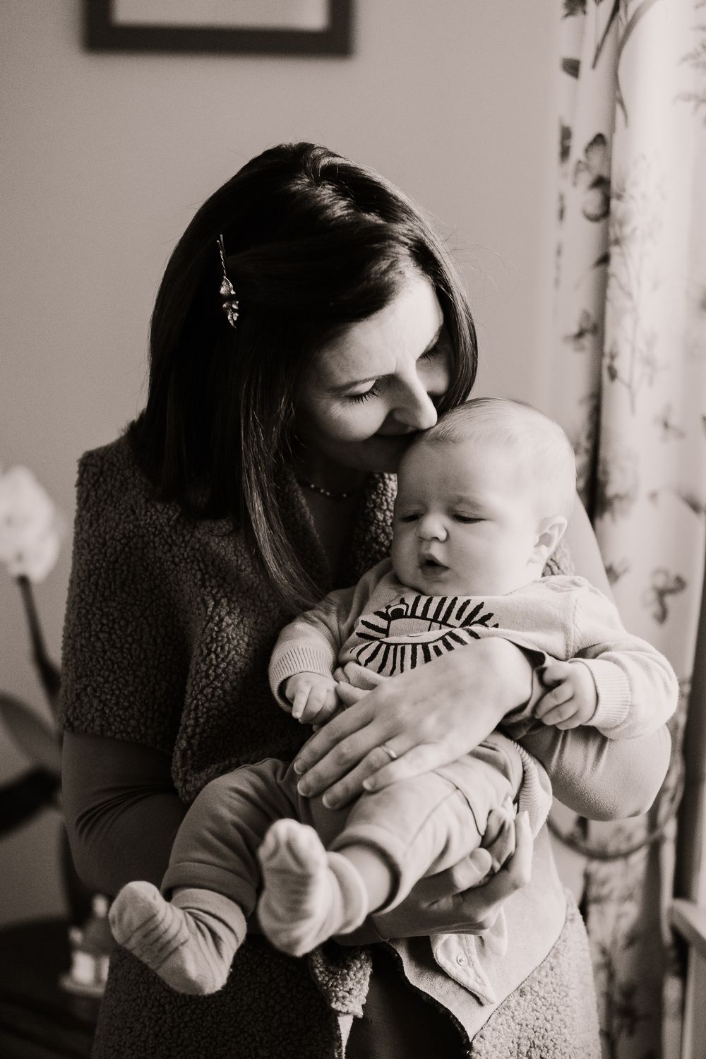 A tender black and white portrait of a parent holding a sleepy baby in a striped outfit by a window.