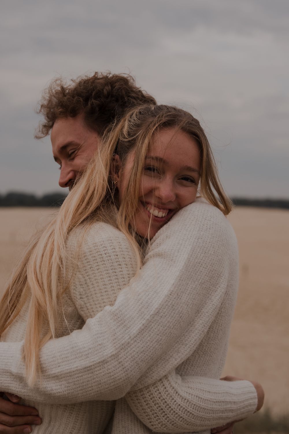 Two people in cozy white sweaters share a warm embrace and laugh together on a cloudy day in an outdoor setting.