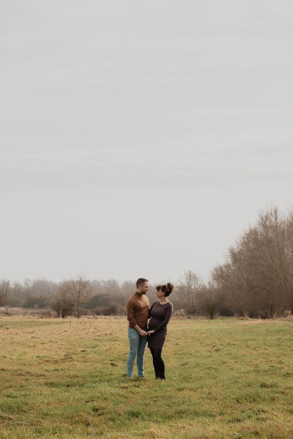 A couple stands together in a misty field surrounded by bare winter trees on an overcast day.