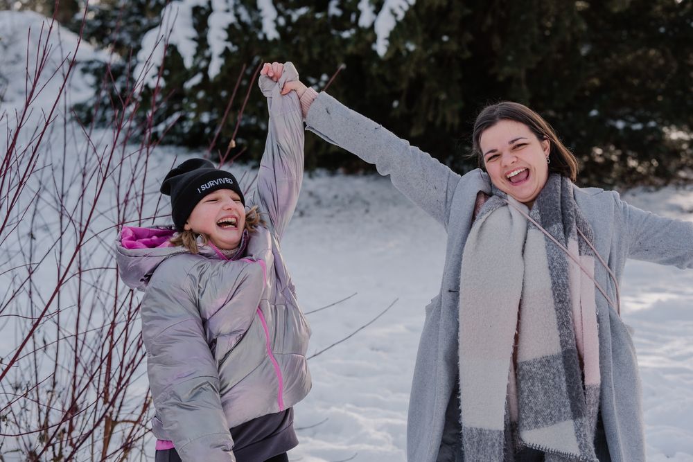 Two people enjoying a snowy winter day outdoors, one jumping excitedly while wearing a lavender coat.