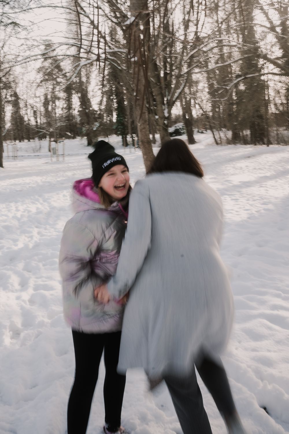 Two people enjoying a winter day in the snow, playfully interacting amid bare trees in a snowy wooded setting.