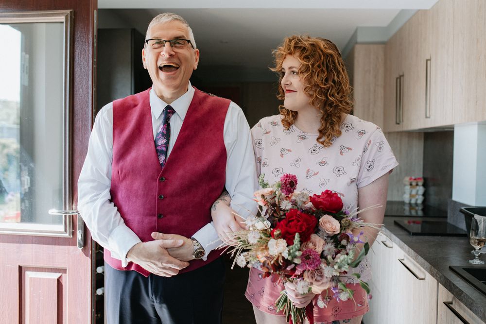 A person wearing a burgundy vest stands next to someone holding a vibrant pink and red floral bouquet in a kitchen.