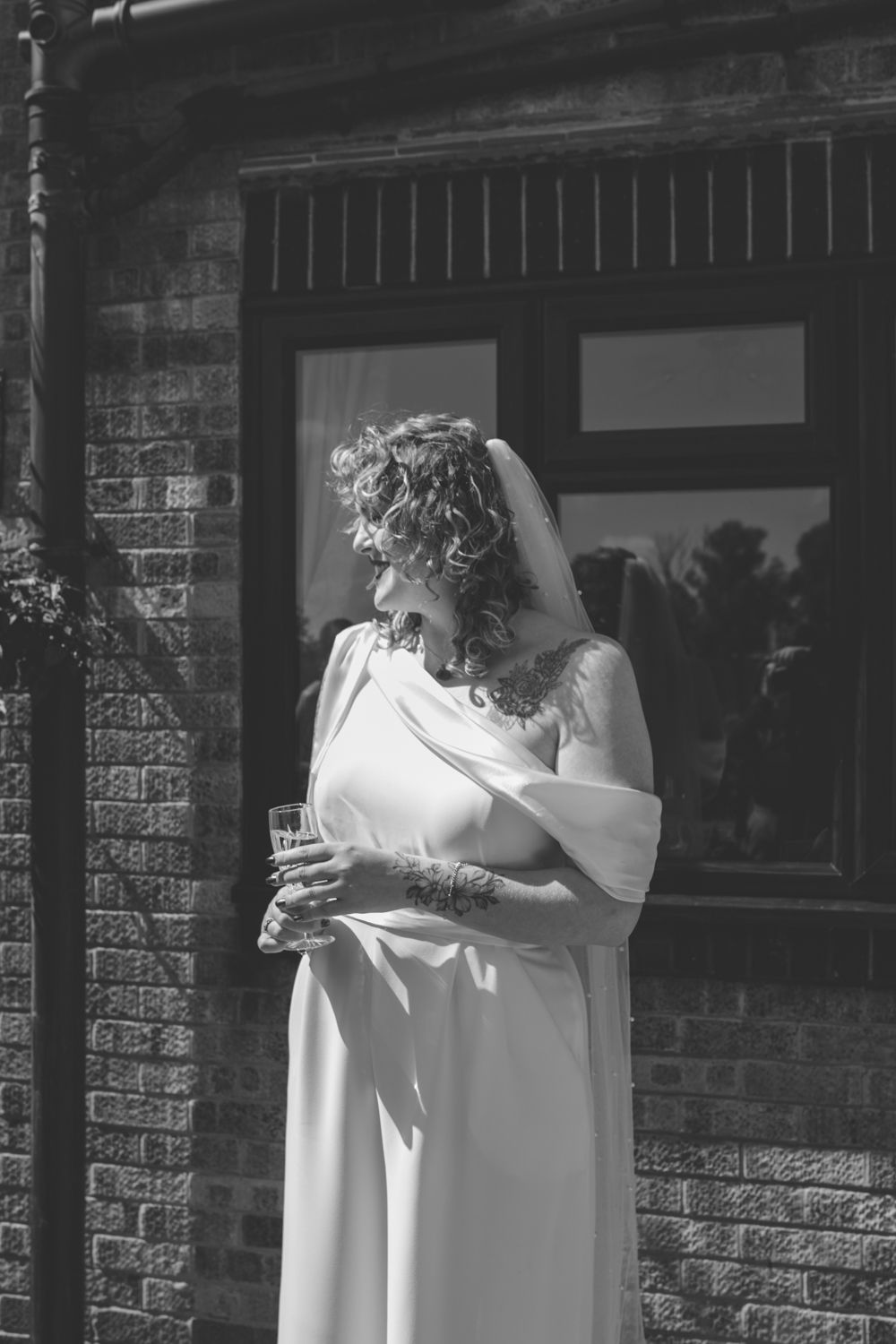 A black and white photograph of someone in a flowing wedding dress standing against a brick wall in sunlight.