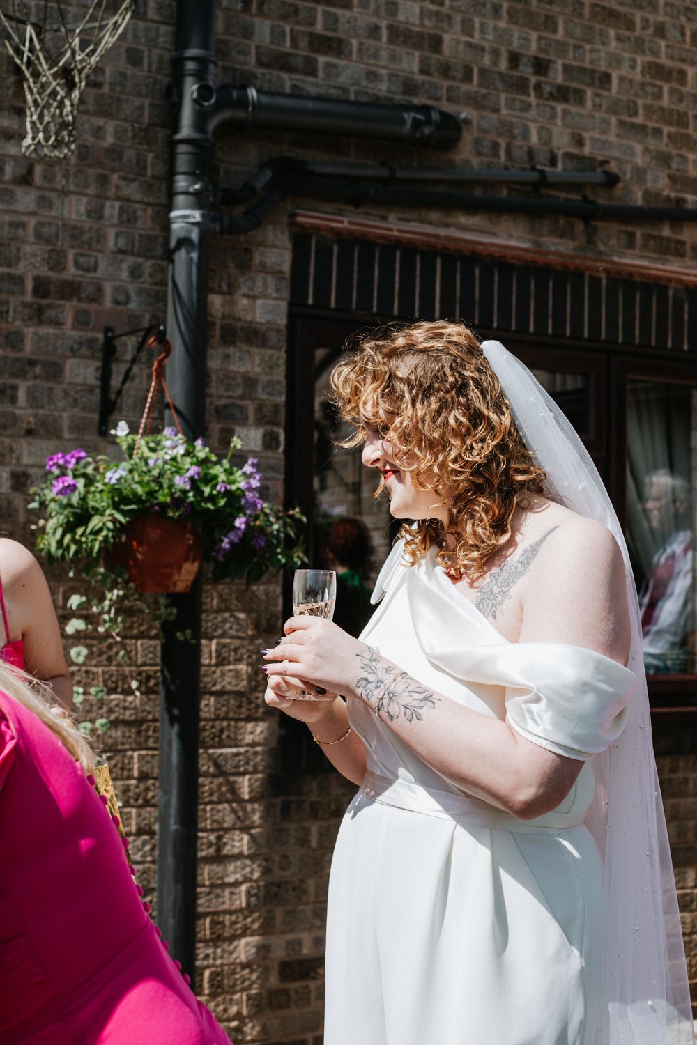 A bride in a white wedding dress and veil holds a glass during an outdoor reception near a brick wall with hanging flowers.