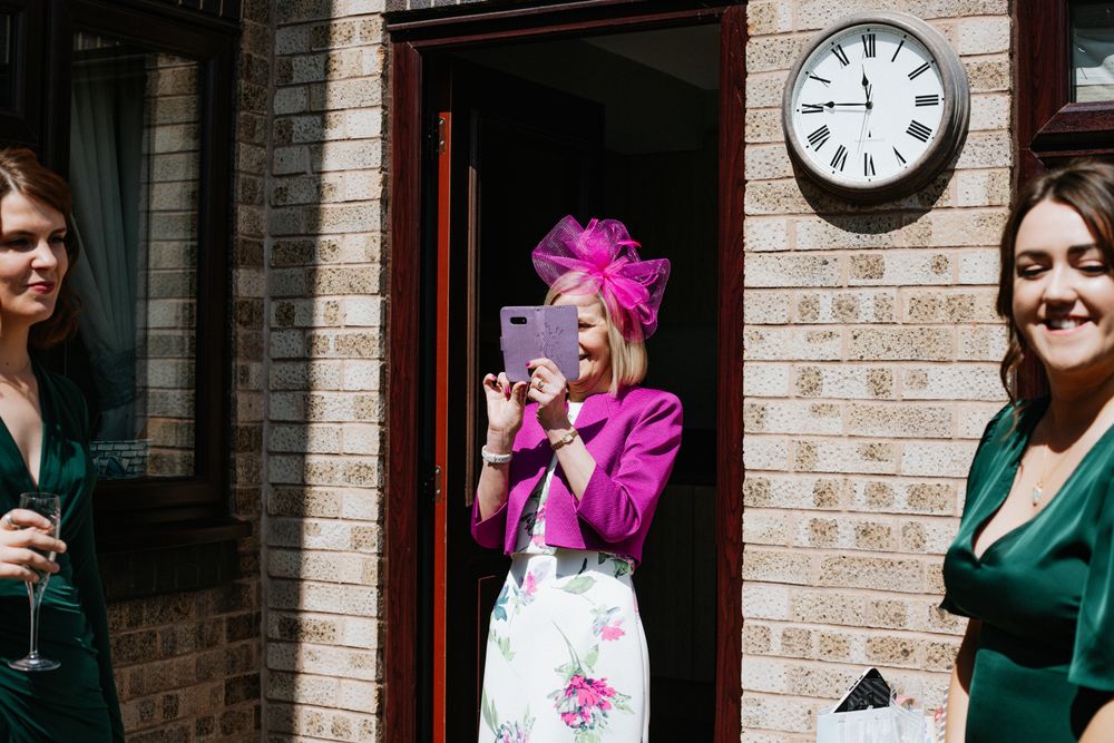 Three people gather at a brick entrance, with one wearing a vibrant pink outfit and fascinator at a formal event.
