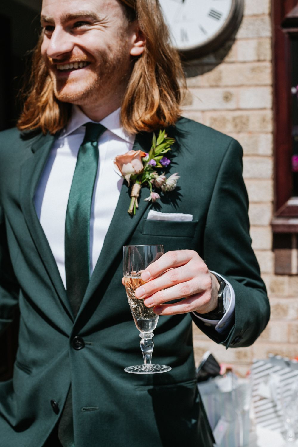 Wedding attendee in dark green suit holds champagne glass while wearing boutonniere flower pin at formal event.