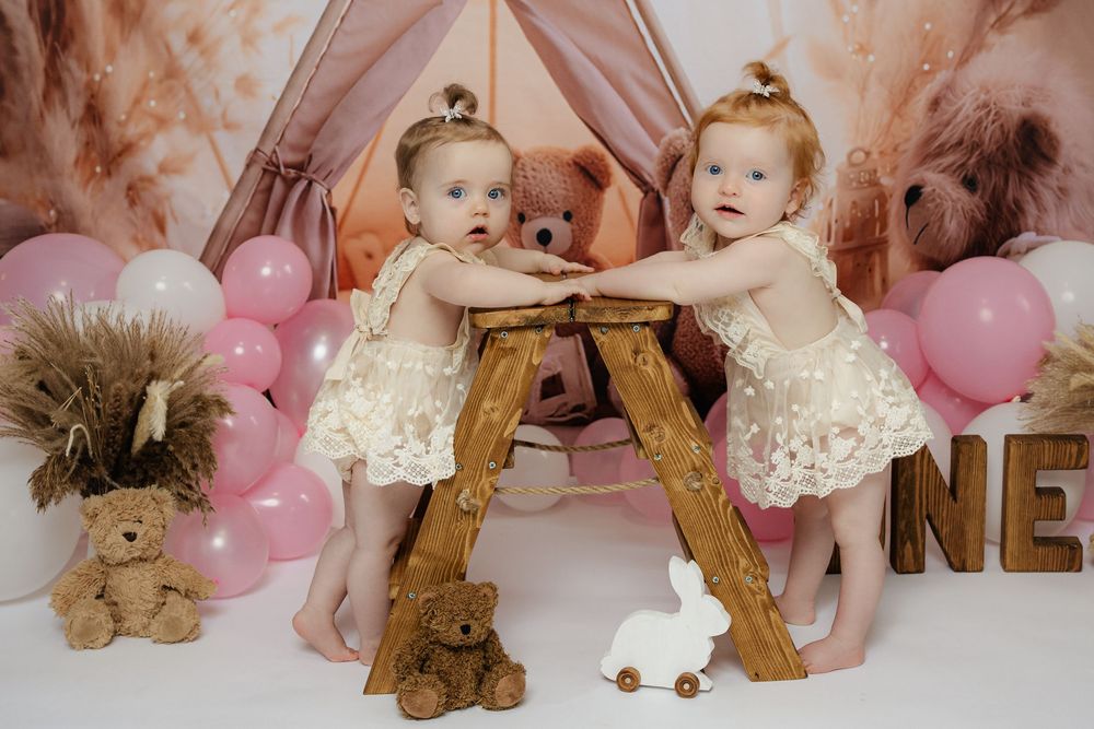 Two babies in white lace dresses stand at wooden ladder with pink balloons and teddy bear decorations in background.