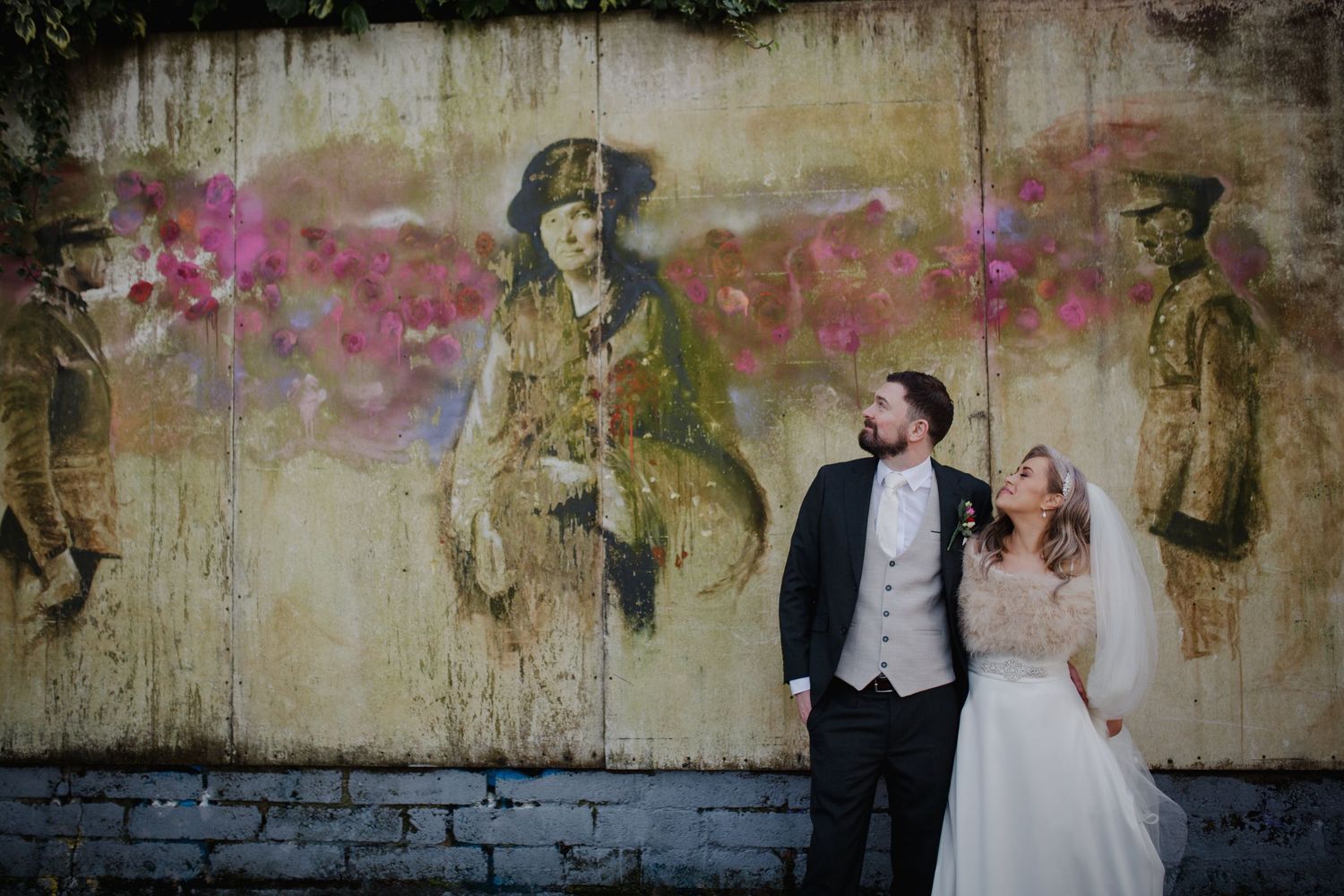 Wedding couple posing against weathered wall with vintage military figure murals and pink floral accents.