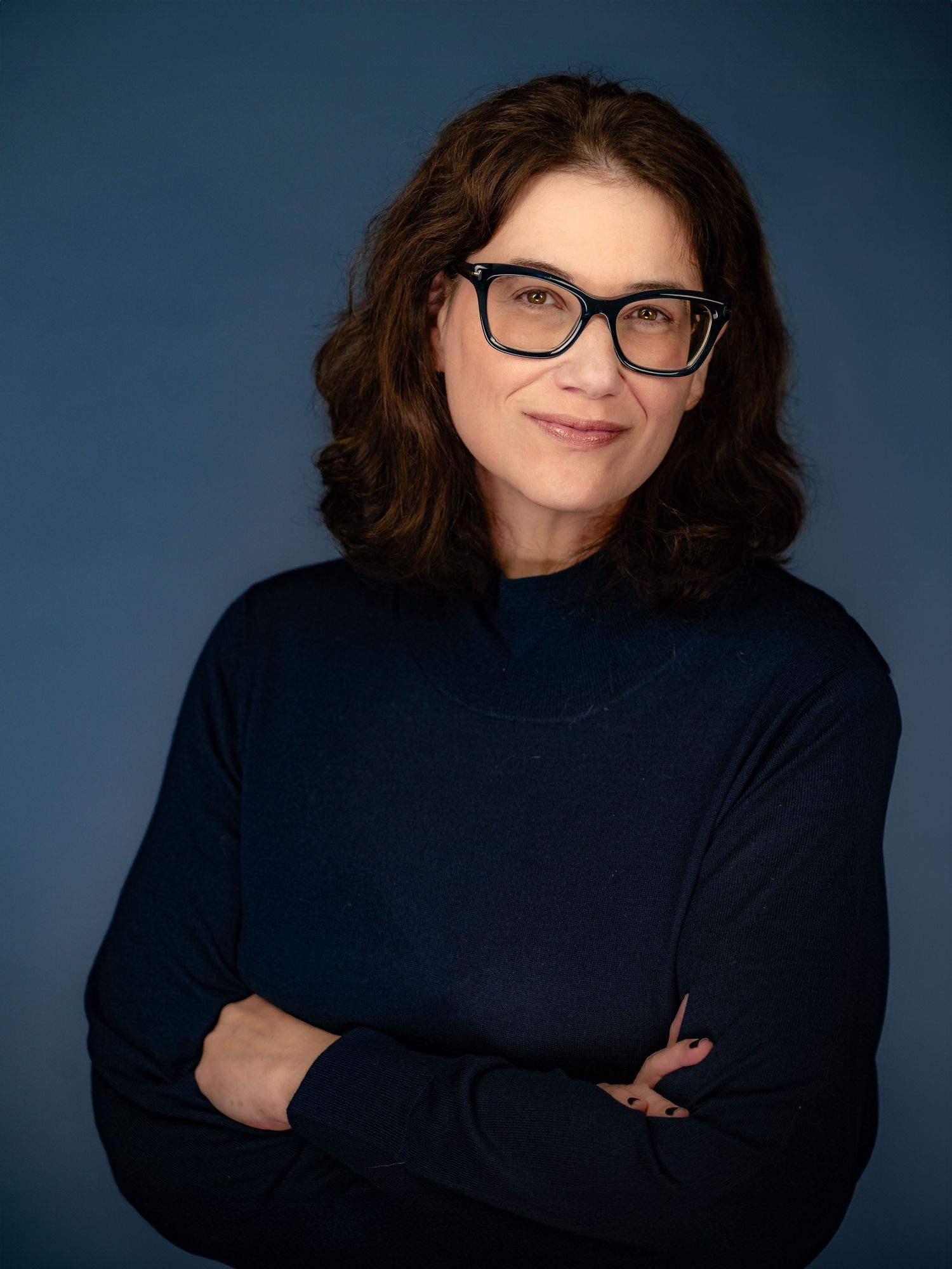 Professional portrait against a blue background showing someone with curly dark hair, glasses and a navy turtleneck sweater.