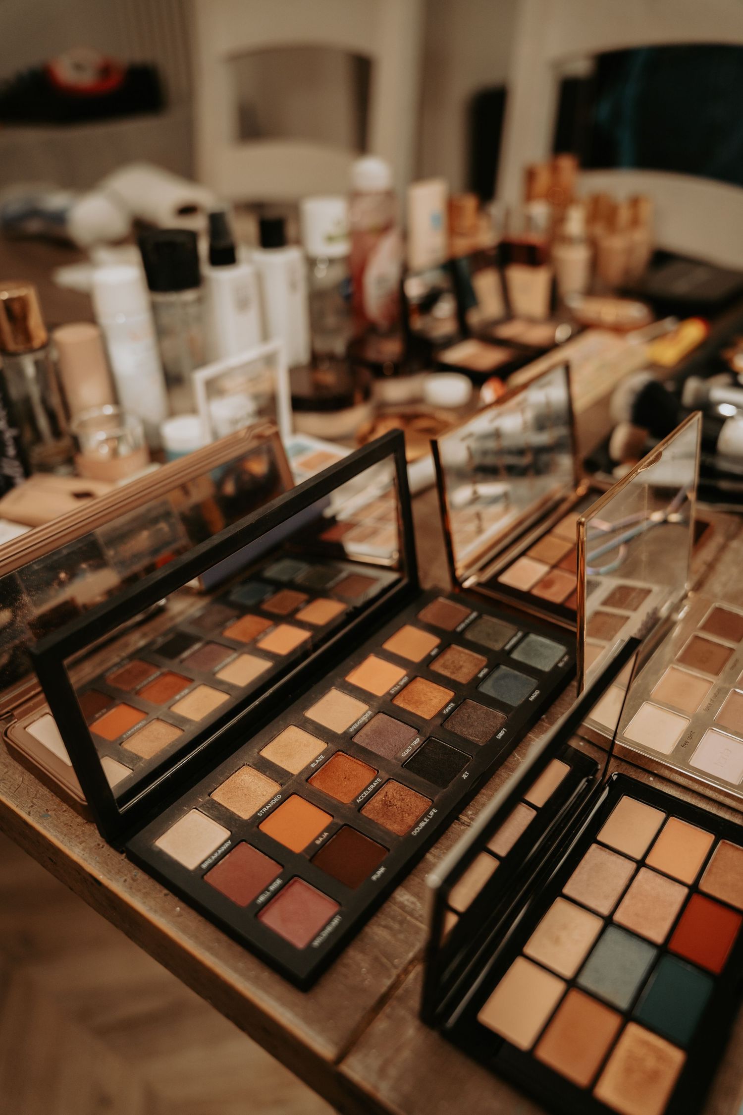 Multiple makeup eyeshadow palettes with neutral and warm tones displayed on a surface with other cosmetic products.