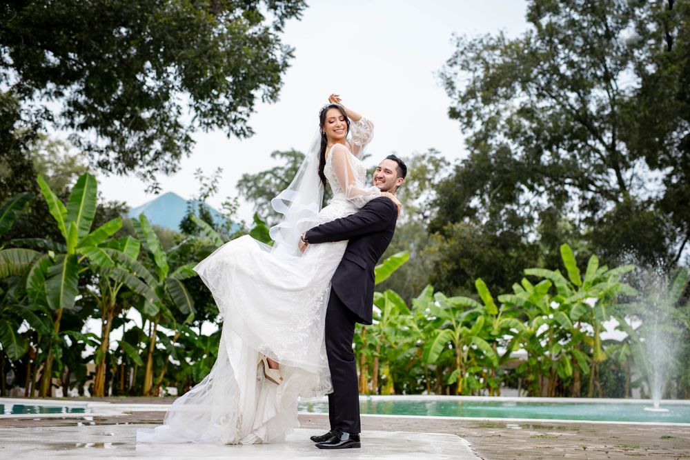 A joyful wedding couple poses outdoors with lush greenery and a fountain in the background.