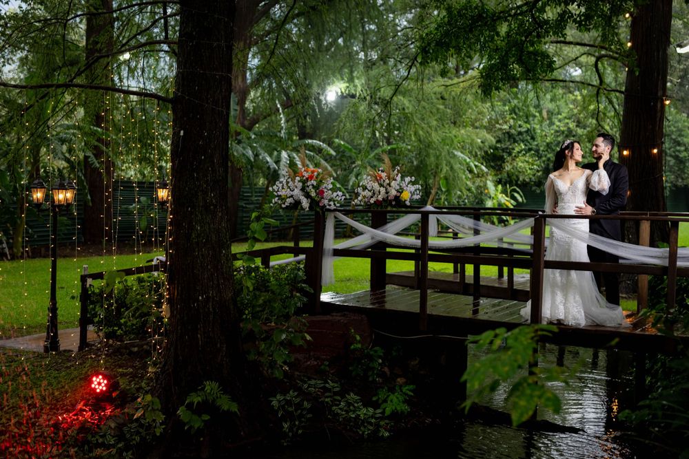 Romantic wedding couple standing on wooden bridge in enchanted forest setting with glowing lights at dusk.