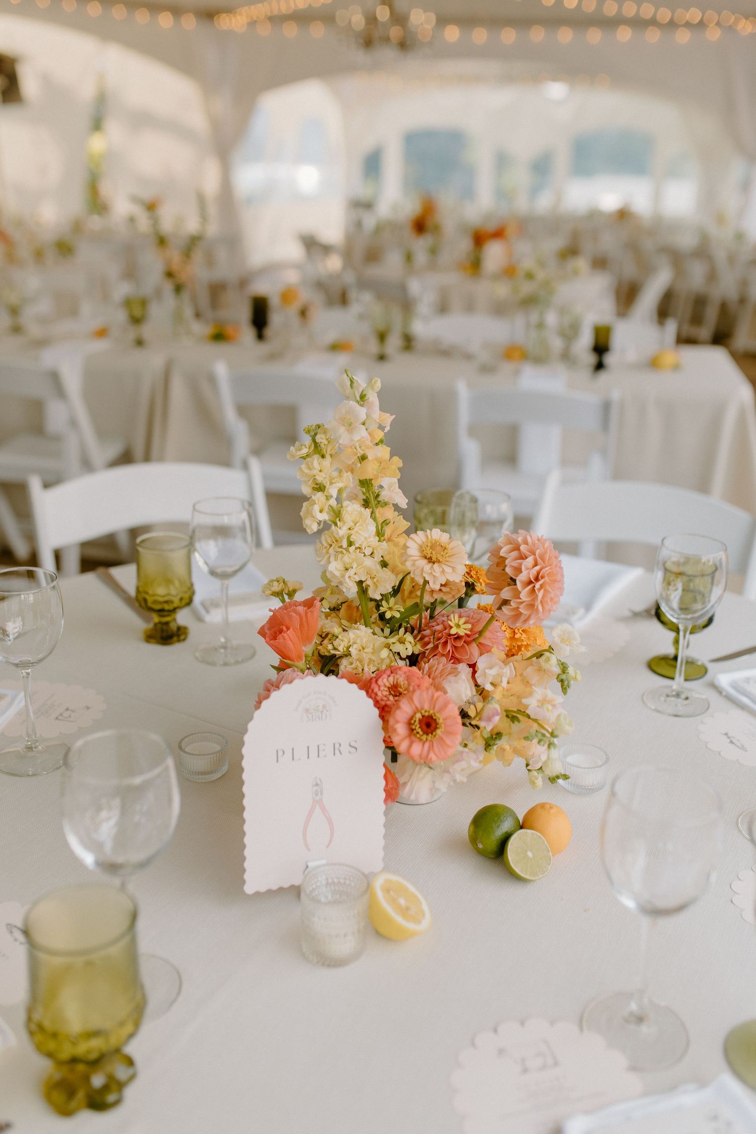 Elegant wedding reception table setting with peach and yellow floral centerpiece and green vintage glassware in white tent venue.