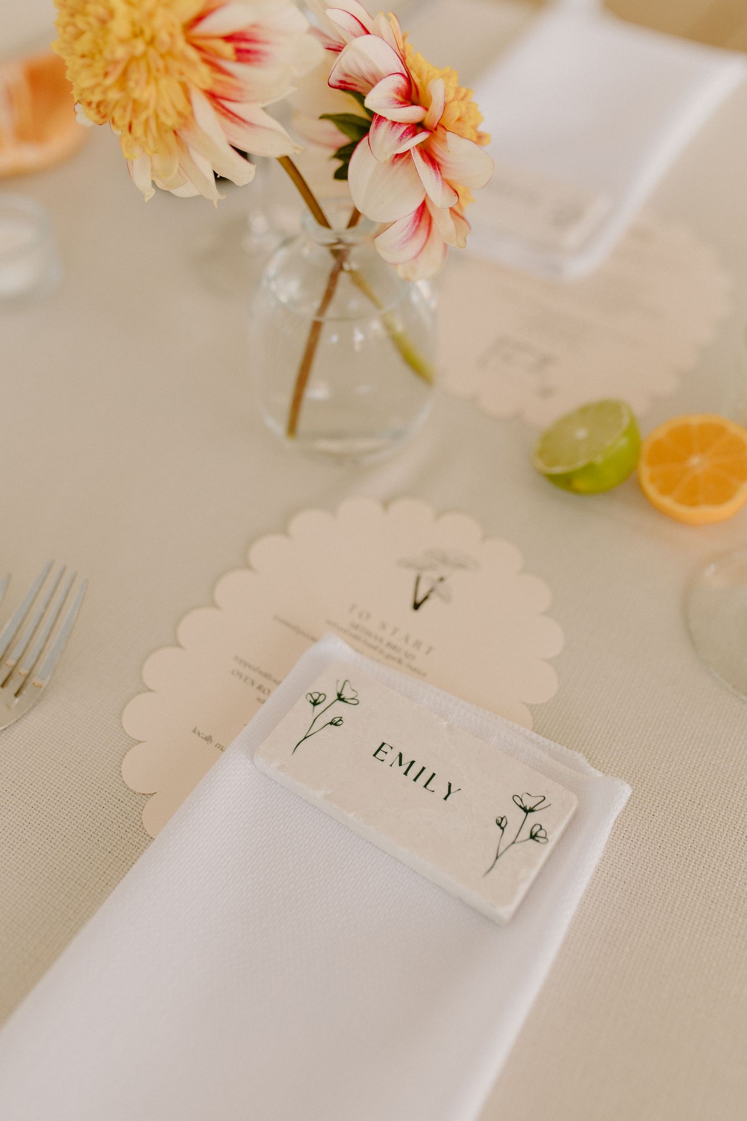 Elegant place setting with floral name card, white napkin, and pink flowers in a glass vase on a cream tablecloth.