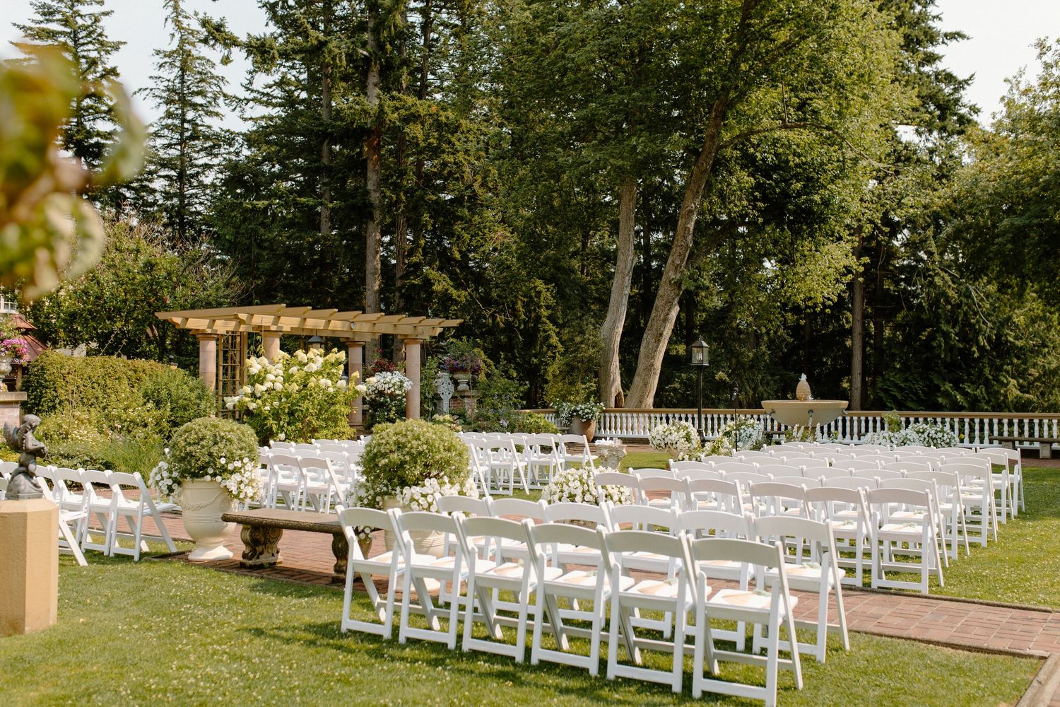 Elegant outdoor wedding ceremony setup with white folding chairs arranged on a green lawn surrounded by tall evergreen trees.