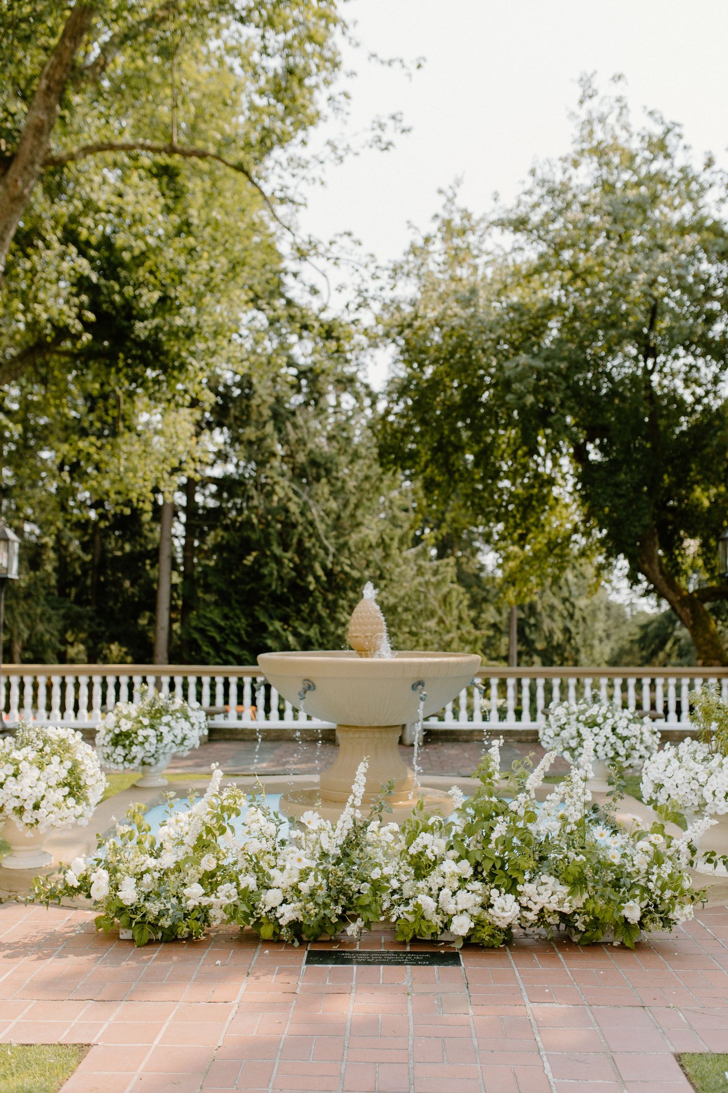 A white classical fountain surrounded by lush white floral arrangements on a terrace with balustrade railing and trees.
