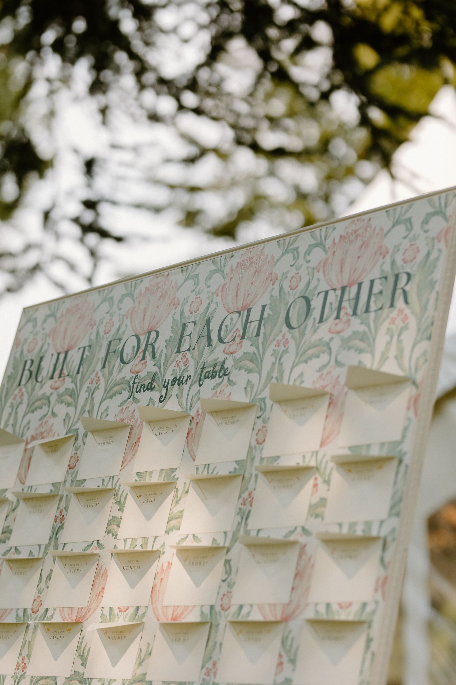 Wedding seating chart board displays 'Built for Each Other' text with arranged name cards in a grid pattern against blurred foliage.