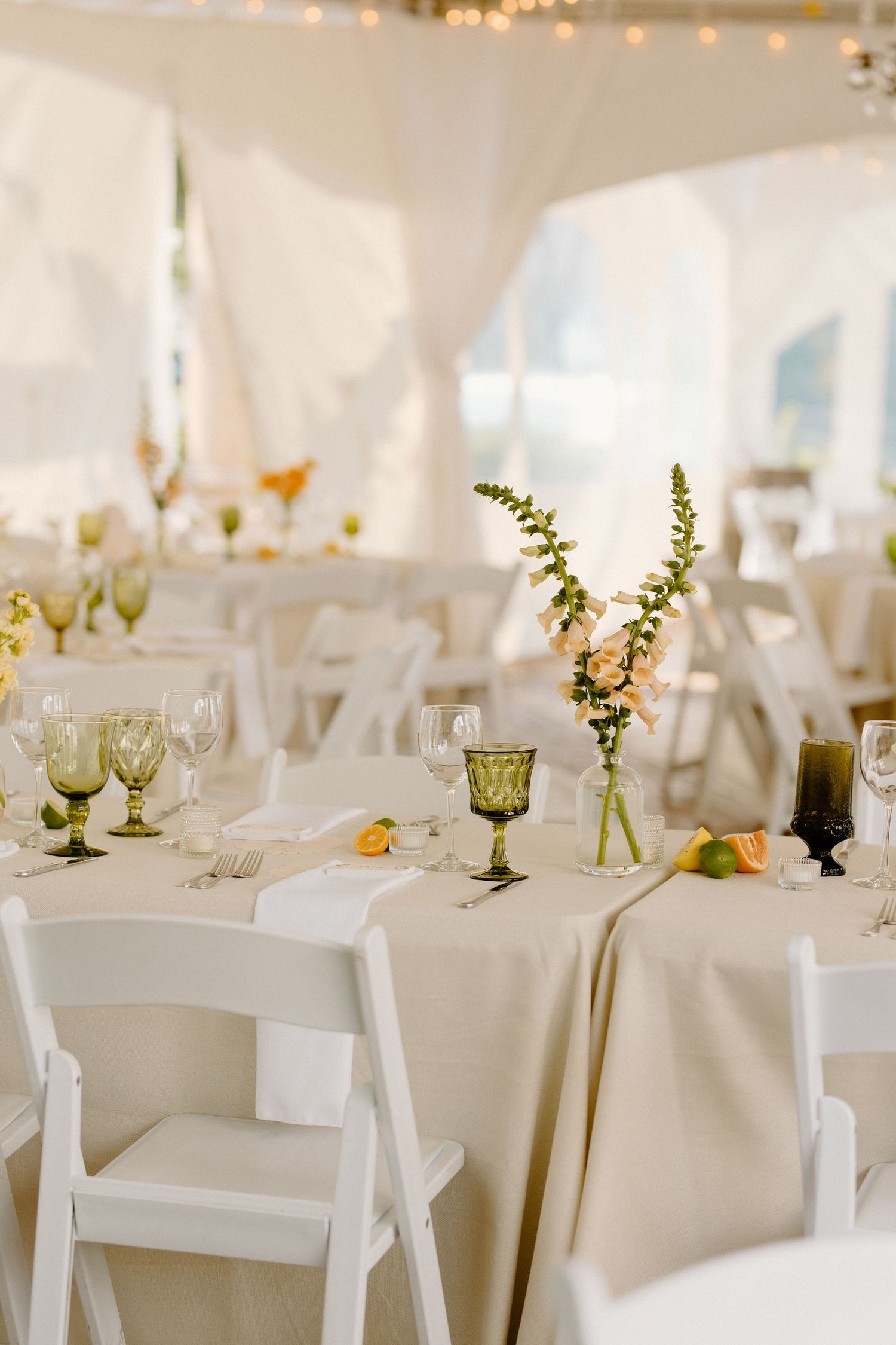 Elegant wedding reception table setting with white linens, green glassware and simple floral centerpieces in a tented venue.