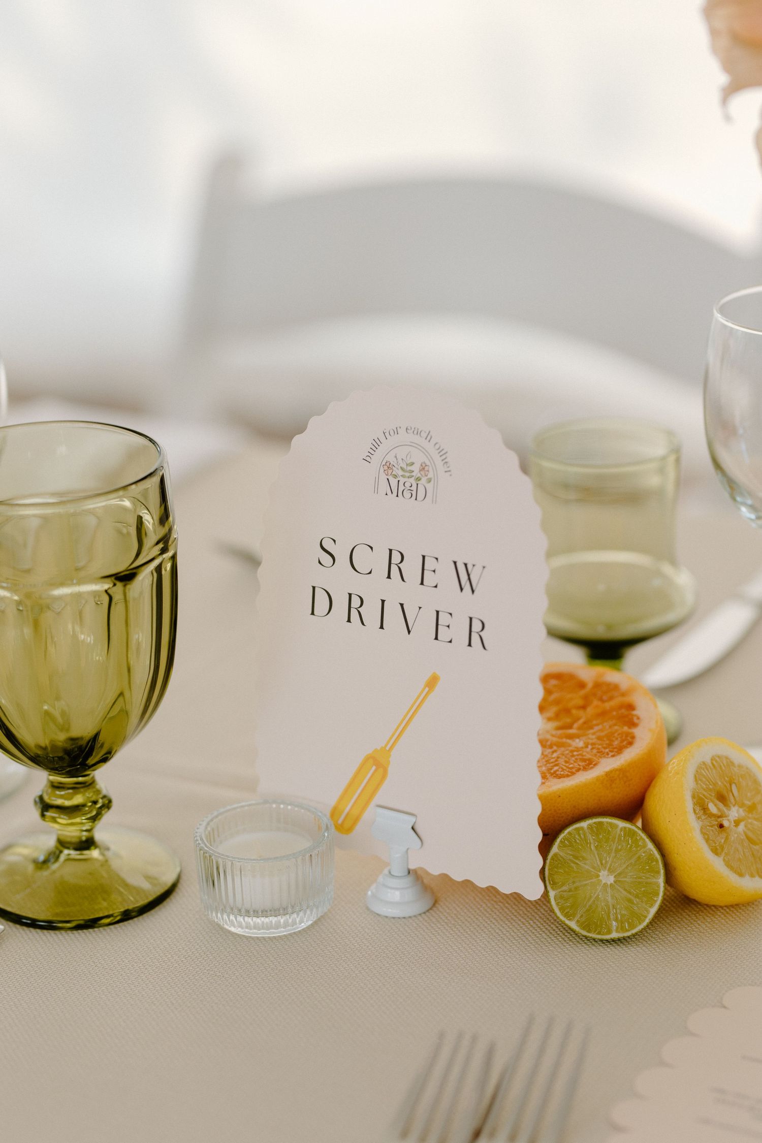 Vintage green glassware and citrus fruits arranged next to a paper cocktail menu titled 'Screwdriver' on a white tablecloth.