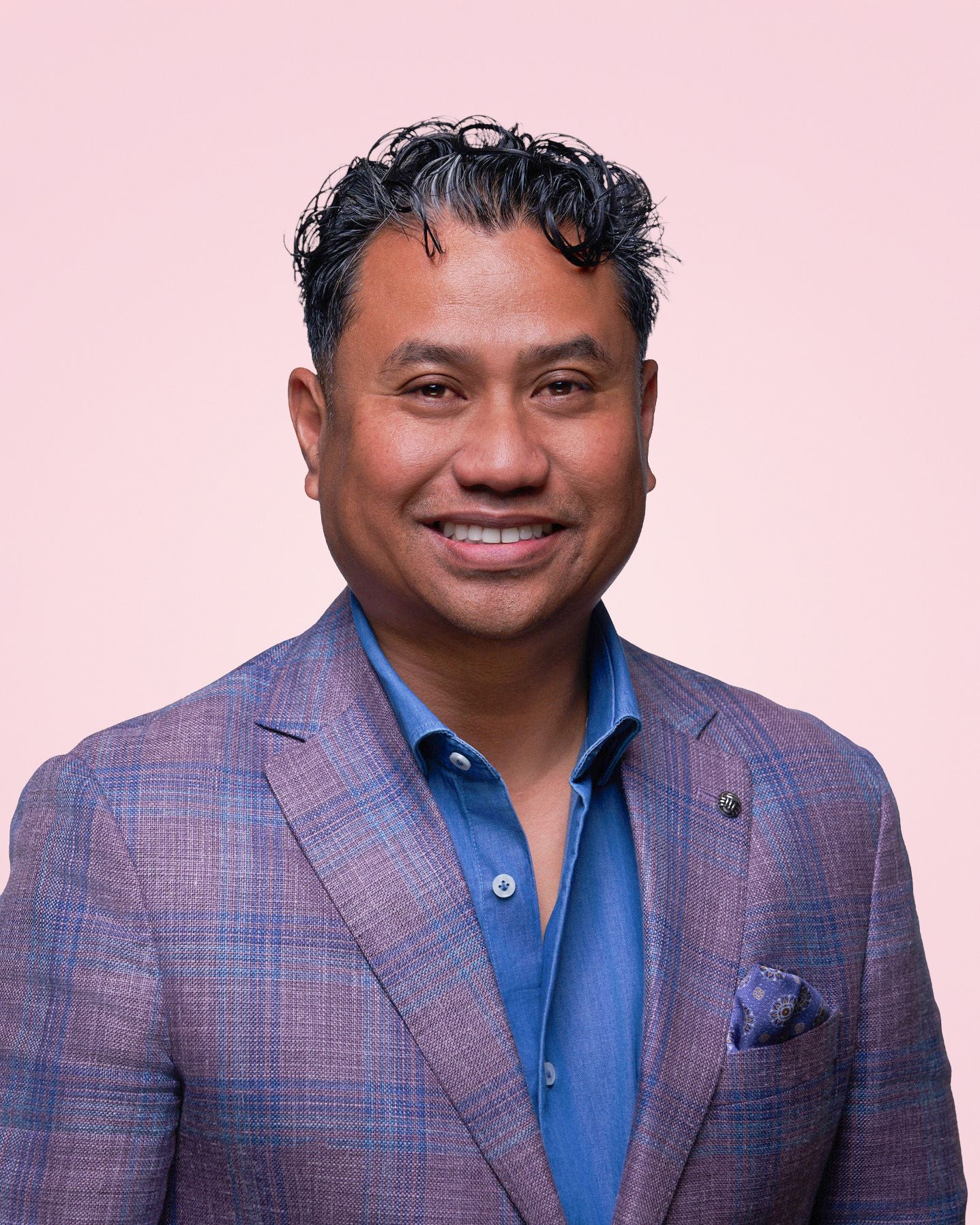 Professional portrait in plaid blazer and blue shirt against pink background.