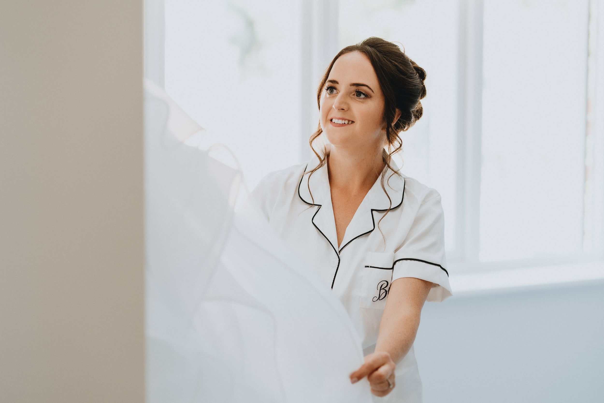 Someone wearing white silk pajamas with black piping smiles warmly in a bright, minimalist setting.