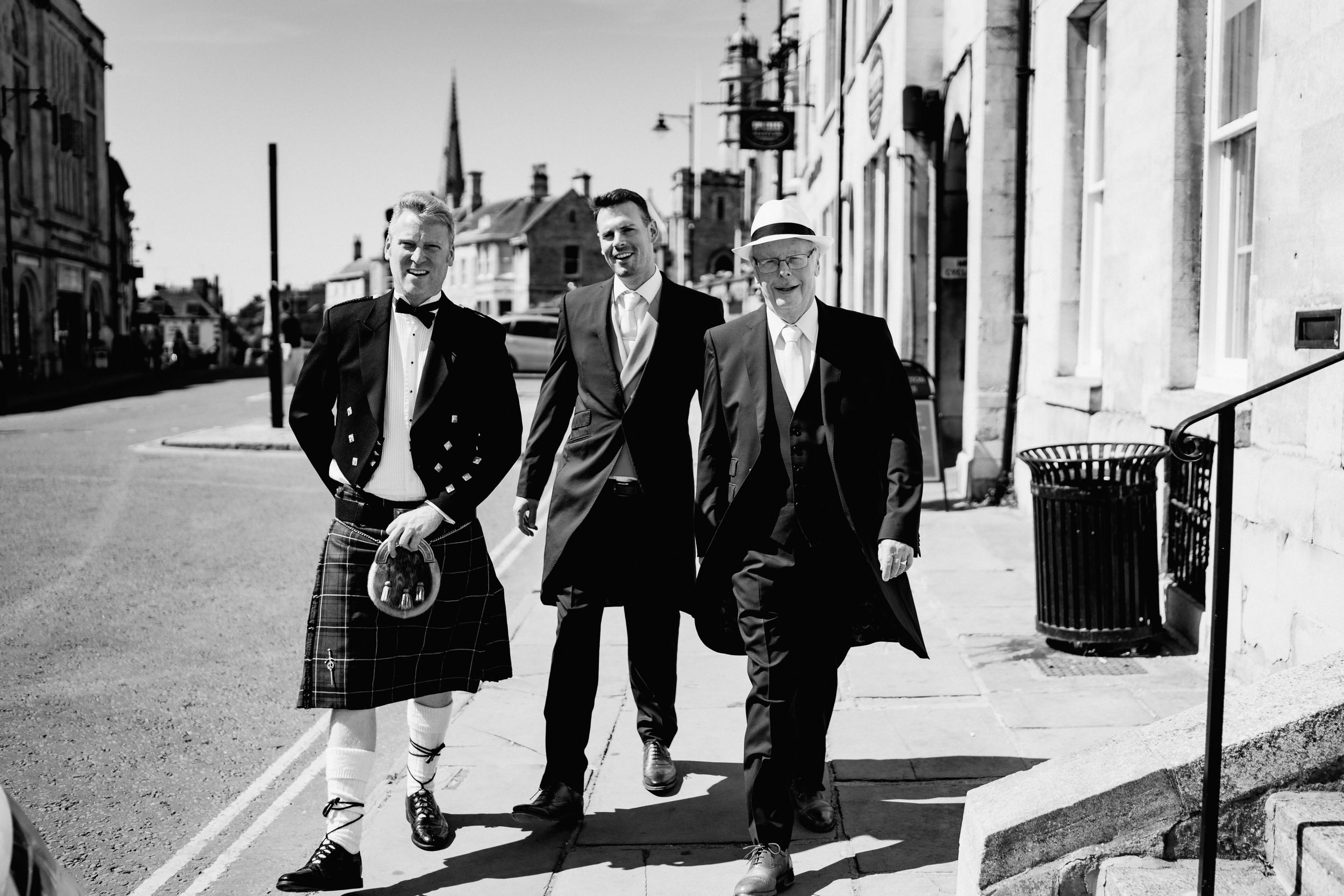 Three men walk together down a Scottish street, with one wearing a traditional kilt and others in dark suits.