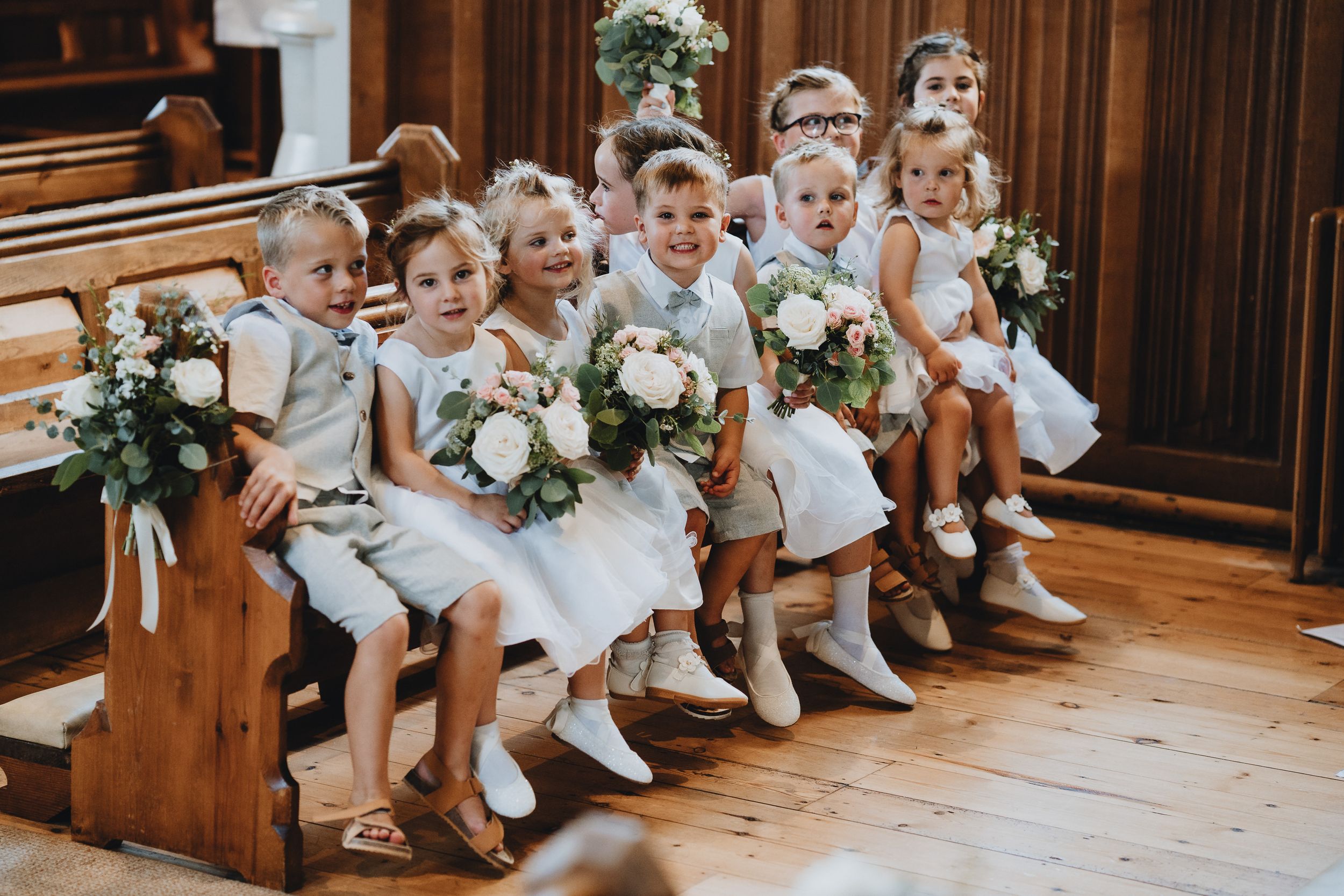 Young attendants in white outfits sit together on a wooden church pew holding floral bouquets.