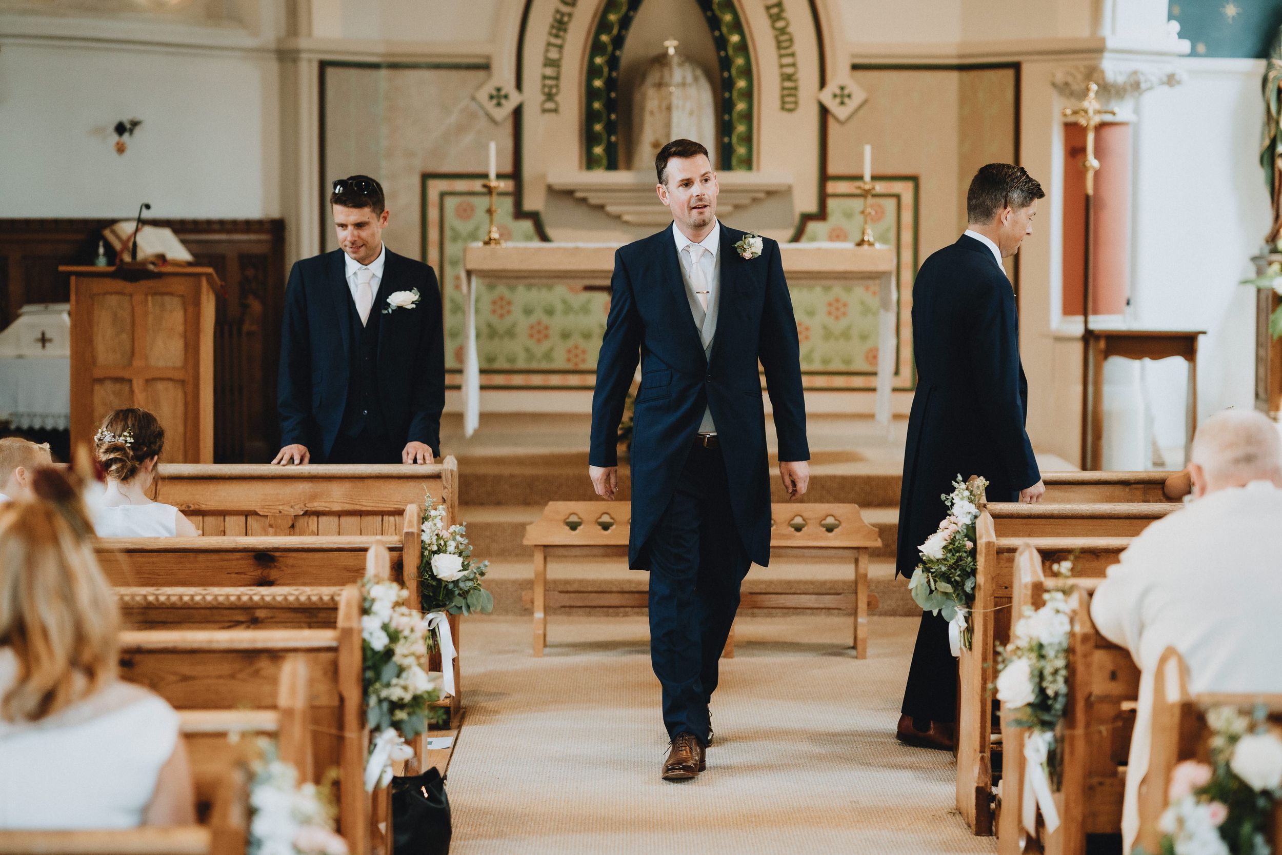 A groomsman walks down the aisle of an ornate church decorated with floral arrangements for a wedding ceremony.