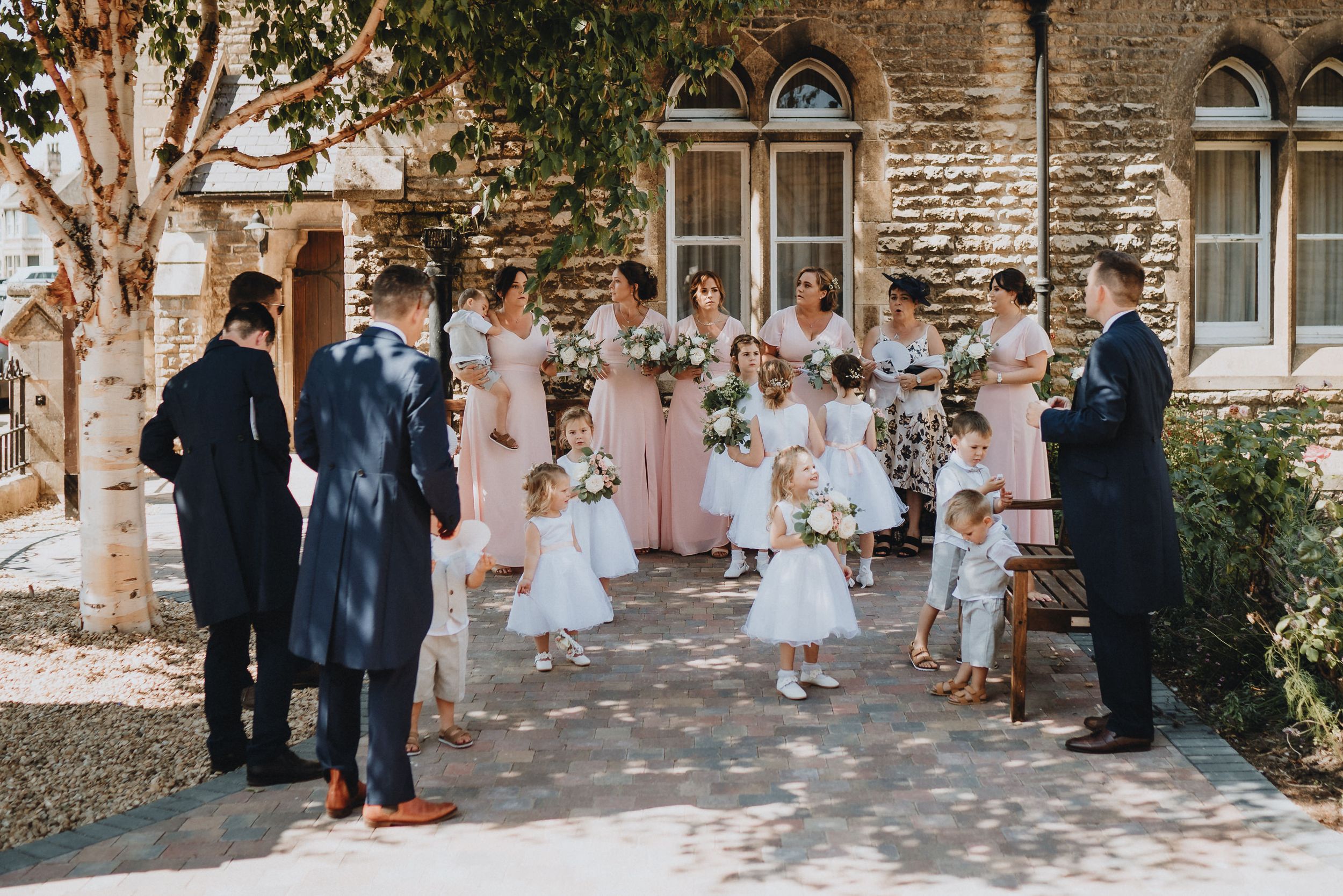 Wedding party gathers on stone pathway outside historic church building with arched windows and greenery.