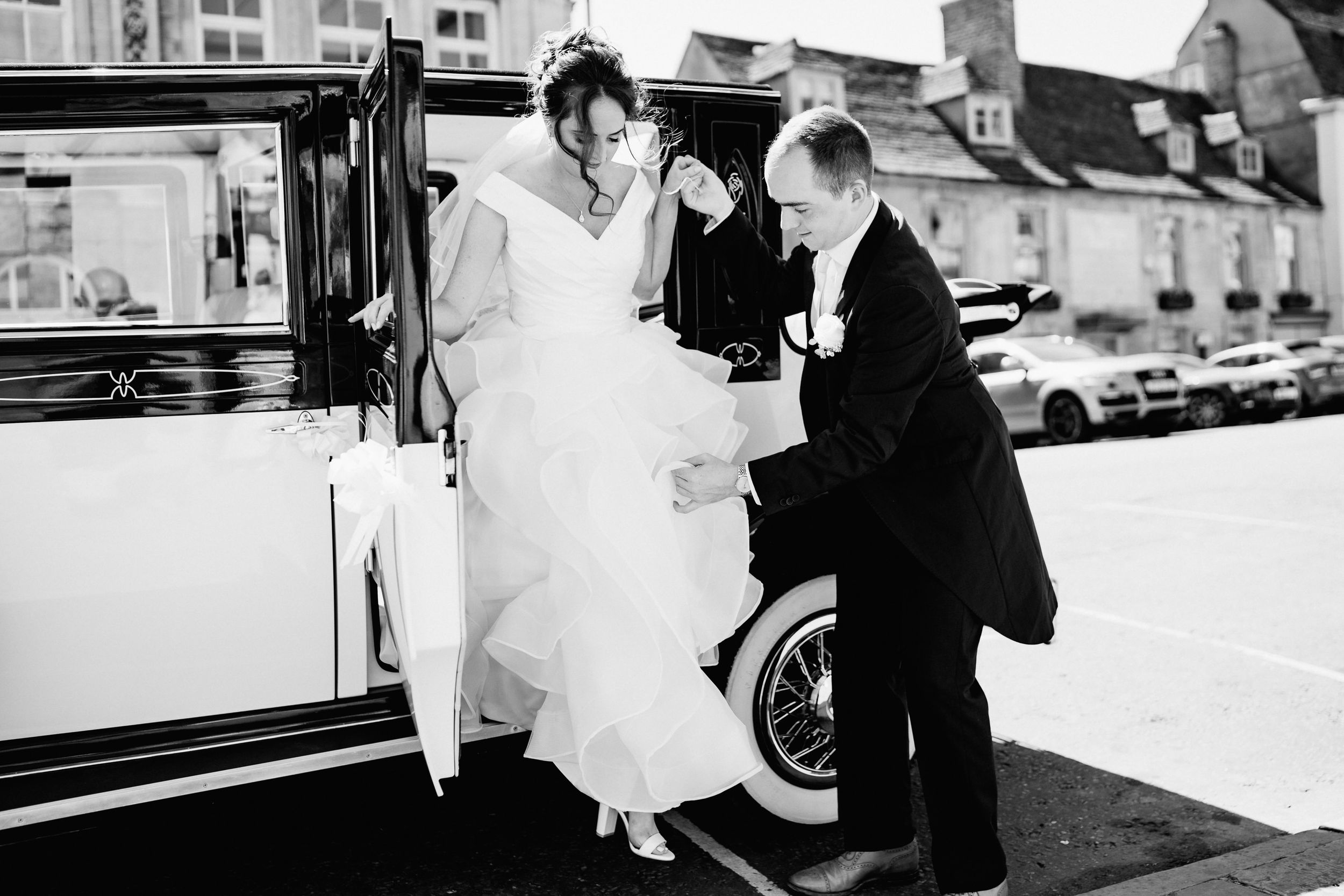 A black and white vintage photo shows a gentleman helping a bride step out of a classic car.