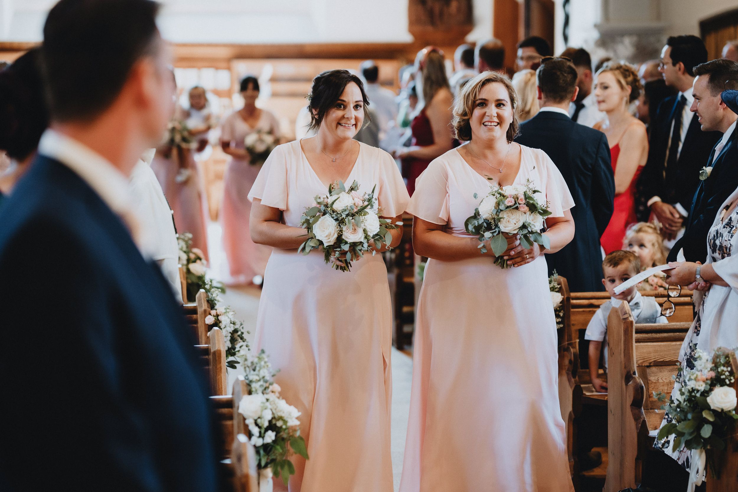 Two bridesmaids in blush pink dresses walk down the church aisle holding white floral bouquets during a wedding ceremony.