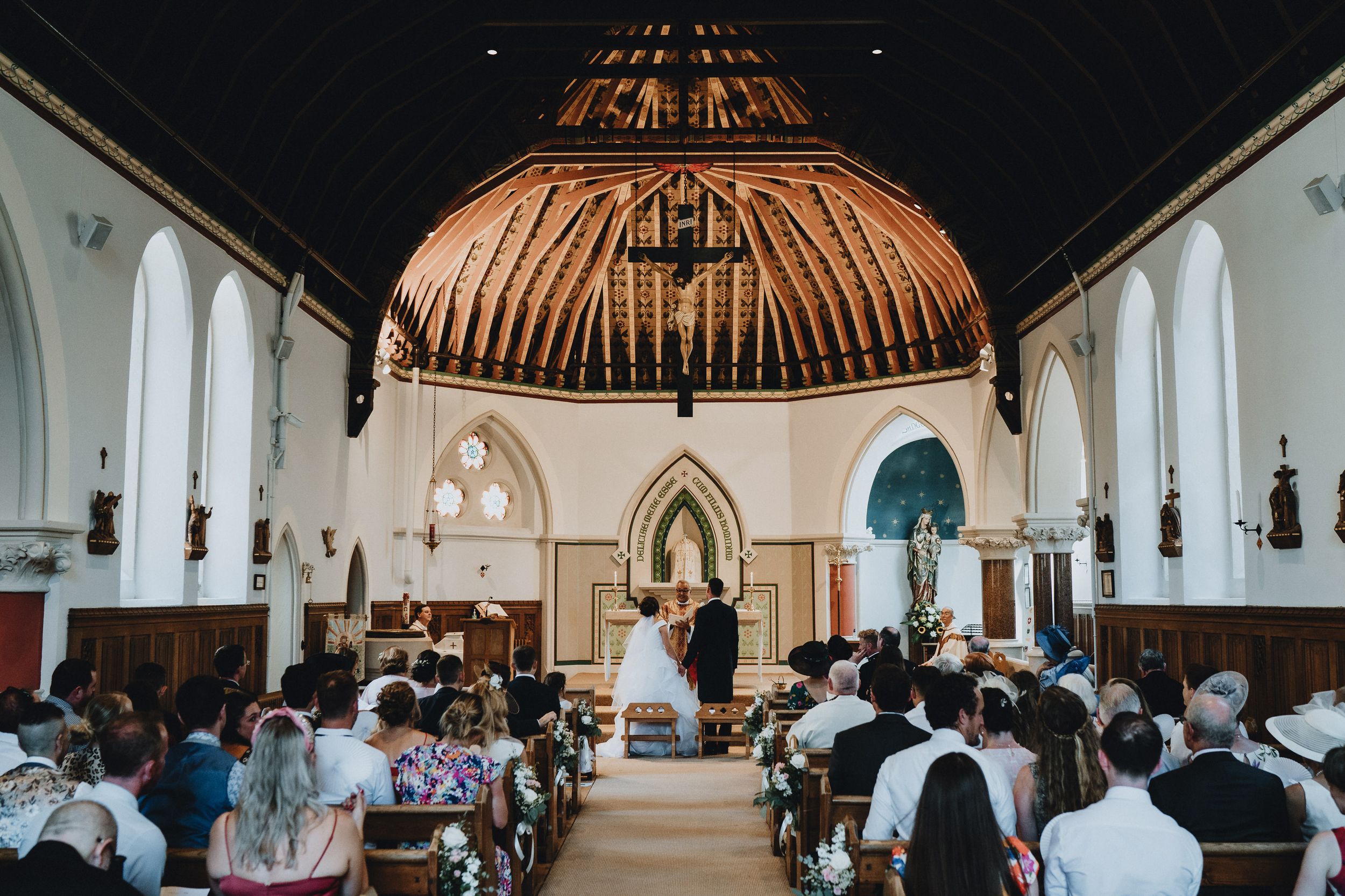 Traditional church wedding ceremony with wooden vaulted ceiling and guests seated in wooden pews watching the ceremony.