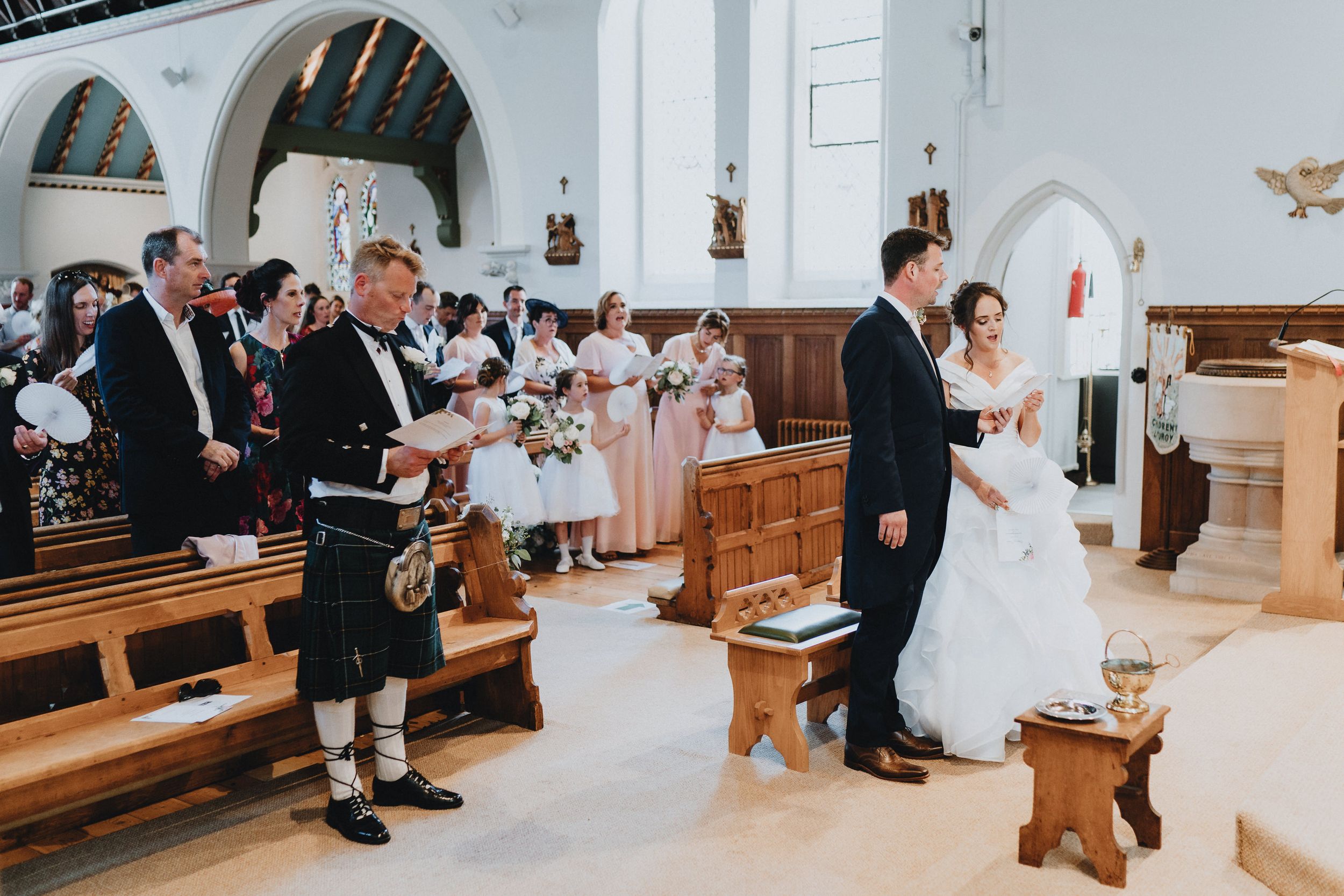Wedding ceremony in a traditional church with a person in a kilt standing among guests in wooden pews.