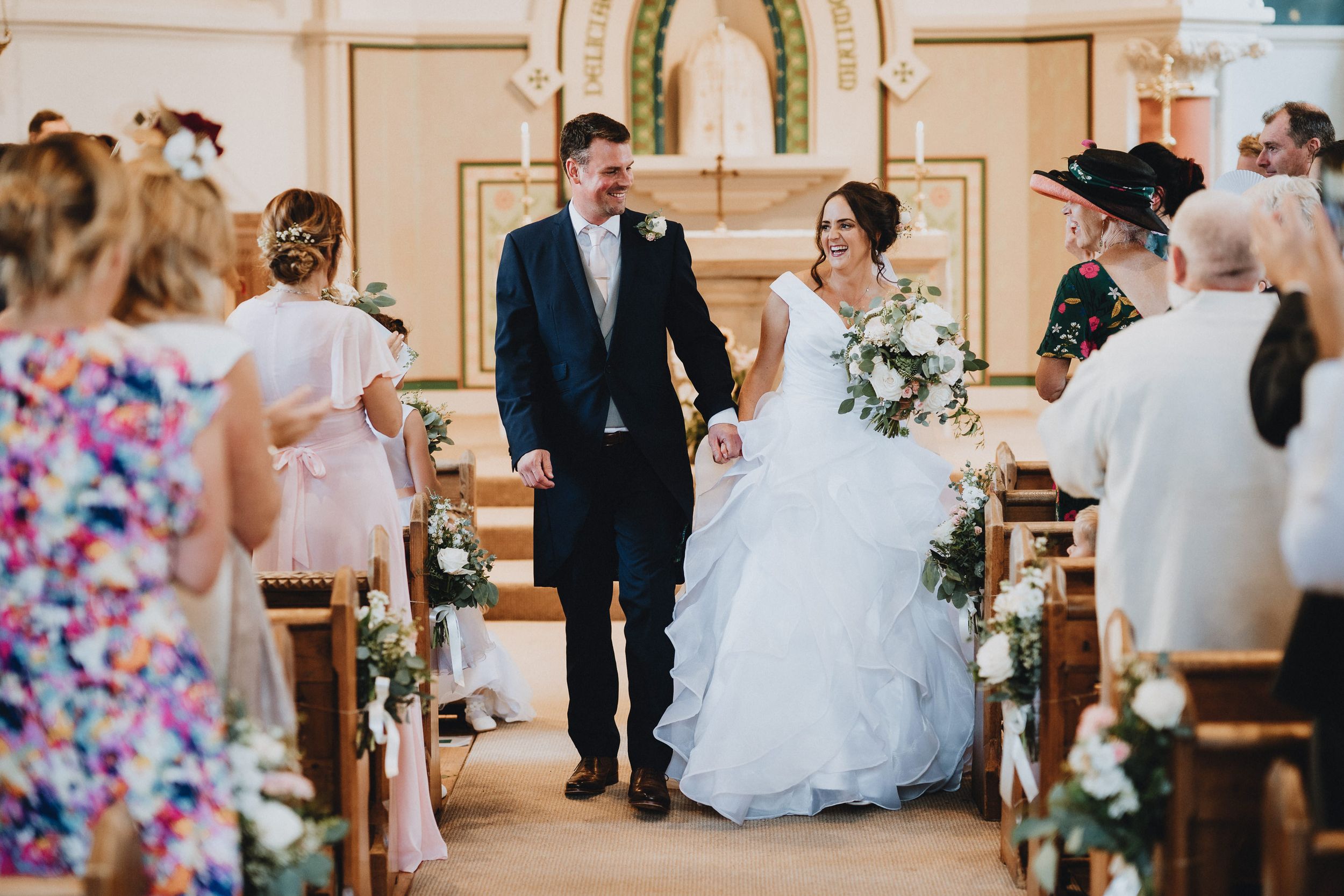 A joyful wedding party walks down the church aisle adorned with floral arrangements.