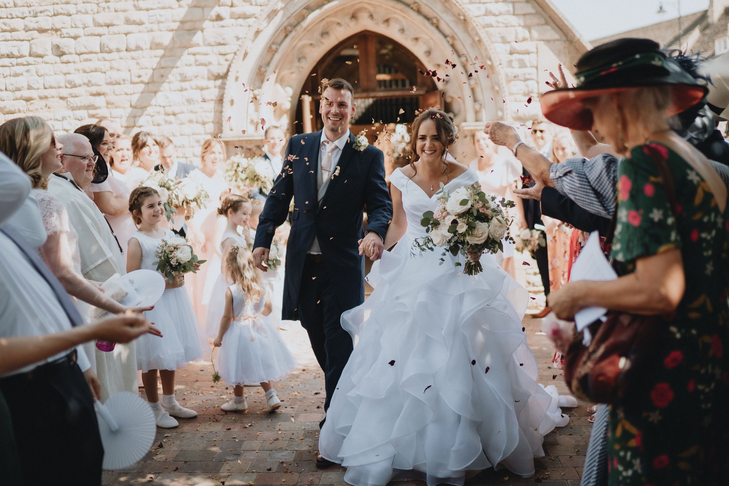 A couple walks through a stone archway at a wedding ceremony while guests shower them with confetti and petals.