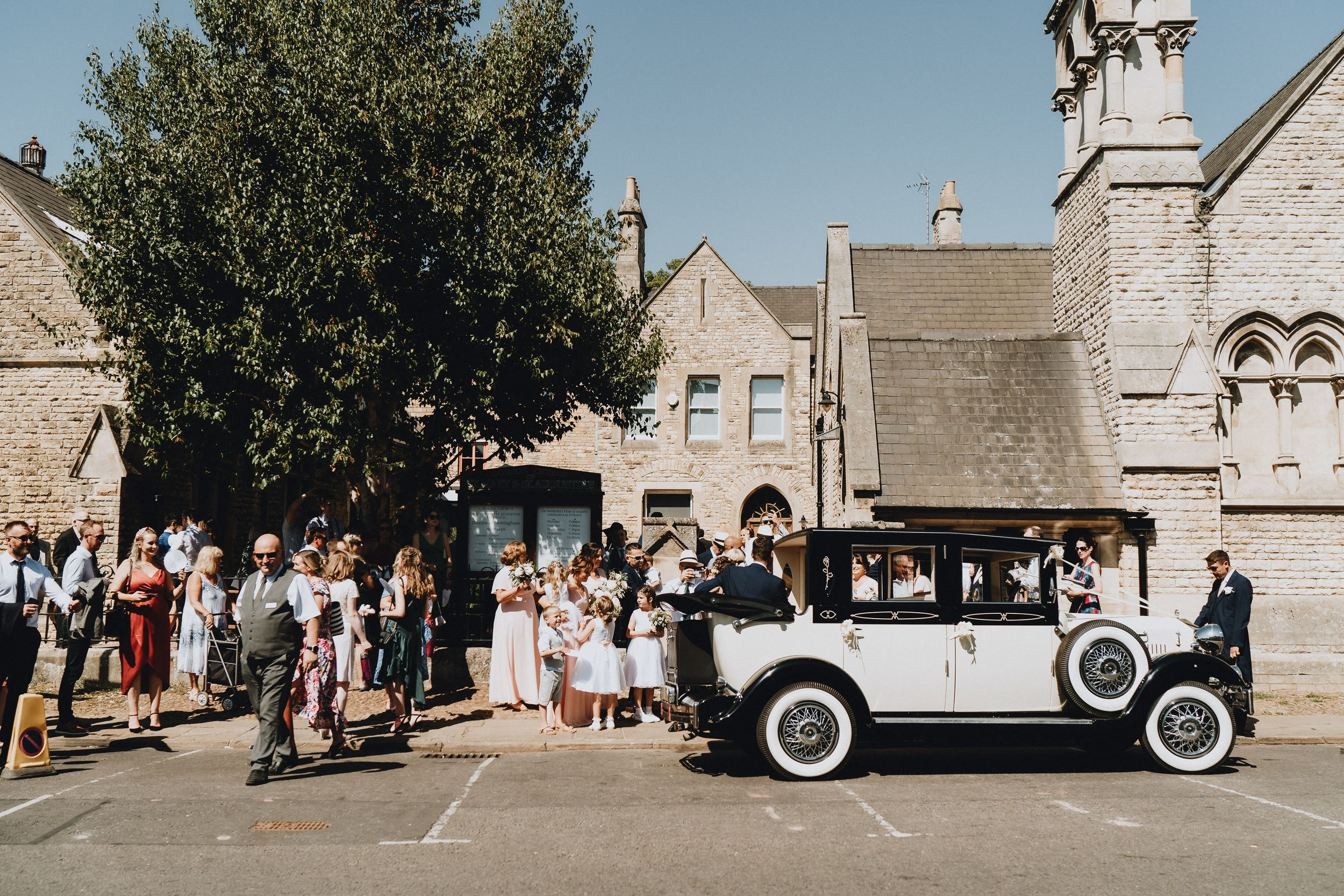 A vintage white classic car parked outside a stone church as a crowd gathers for a special occasion.