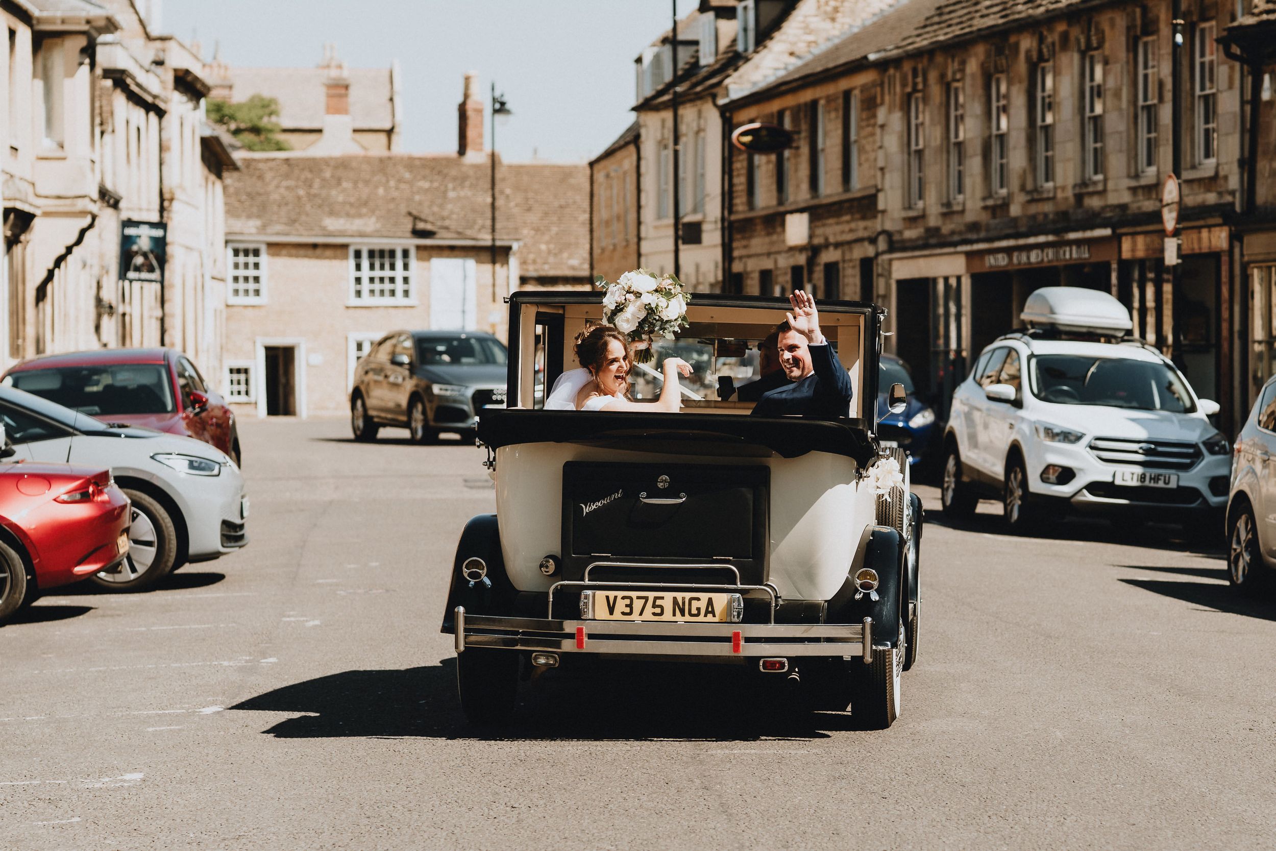 A vintage black car drives down a historic town street lined with stone buildings and modern vehicles.
