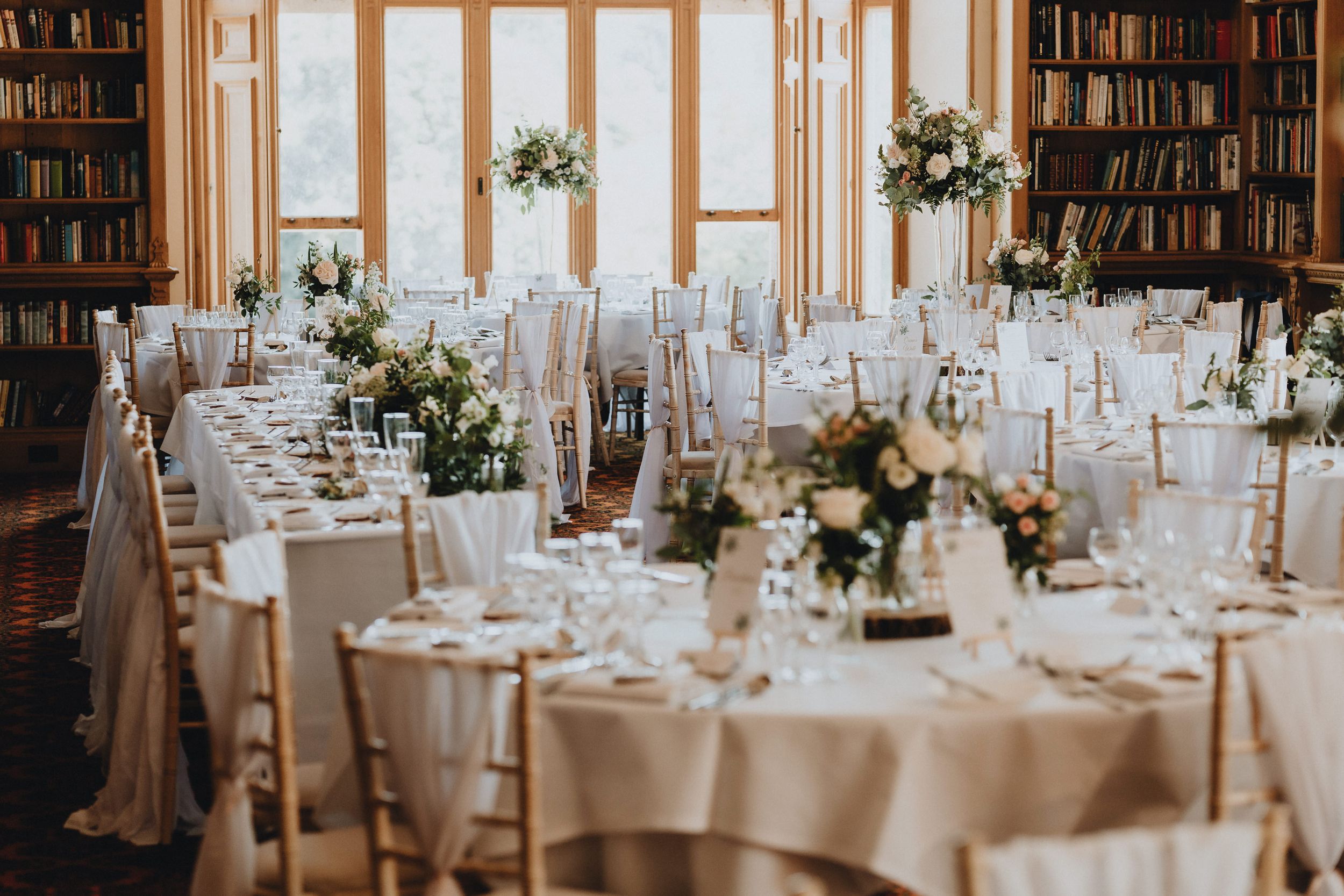Elegant wedding reception tables set with white linens and floral centerpieces in a classic library setting with bookshelves.