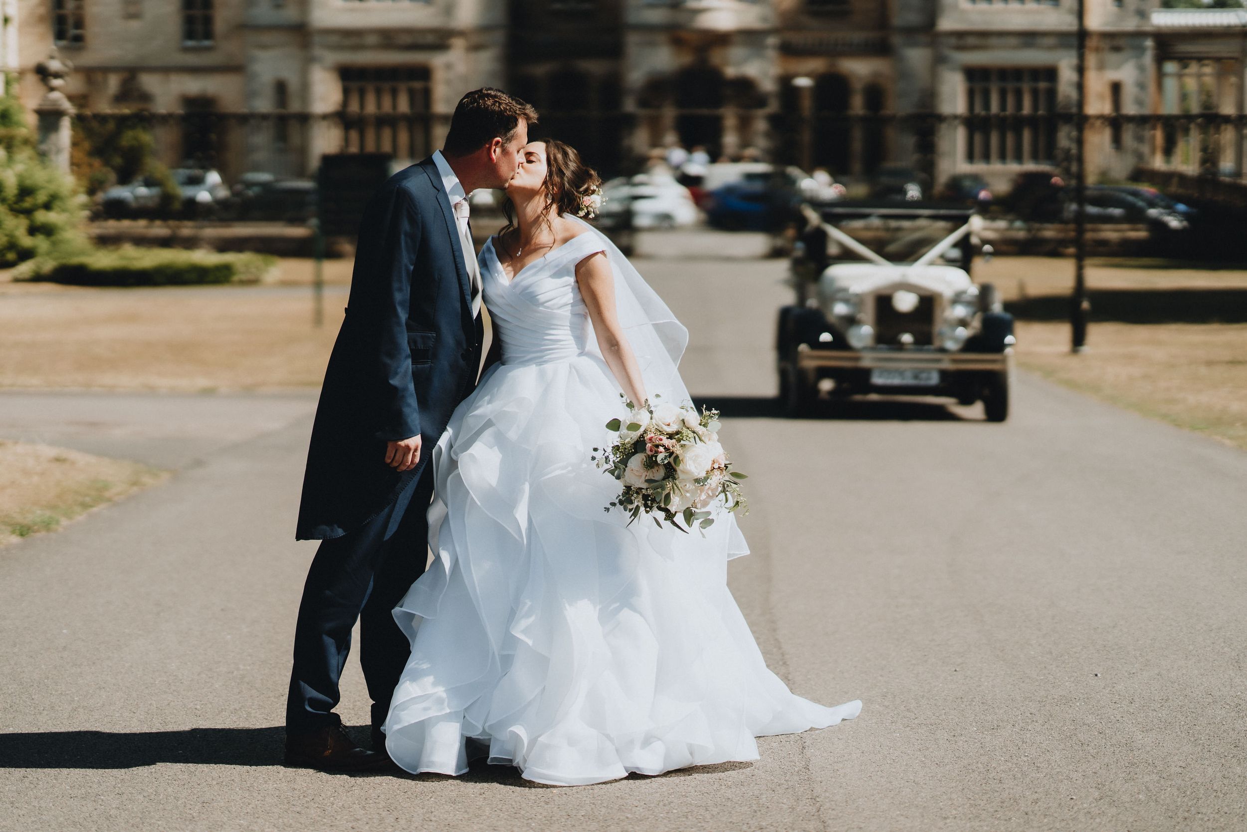 A couple in wedding attire sharing an intimate moment on a driveway with a vintage car in the background.