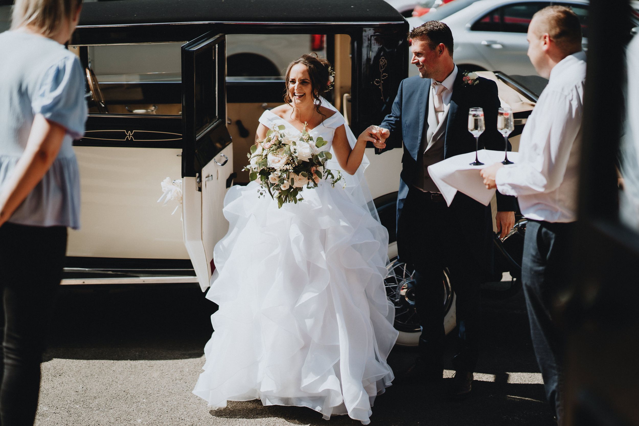 A bride in a flowing white gown is assisted from a vehicle while holding a bouquet on her wedding day.