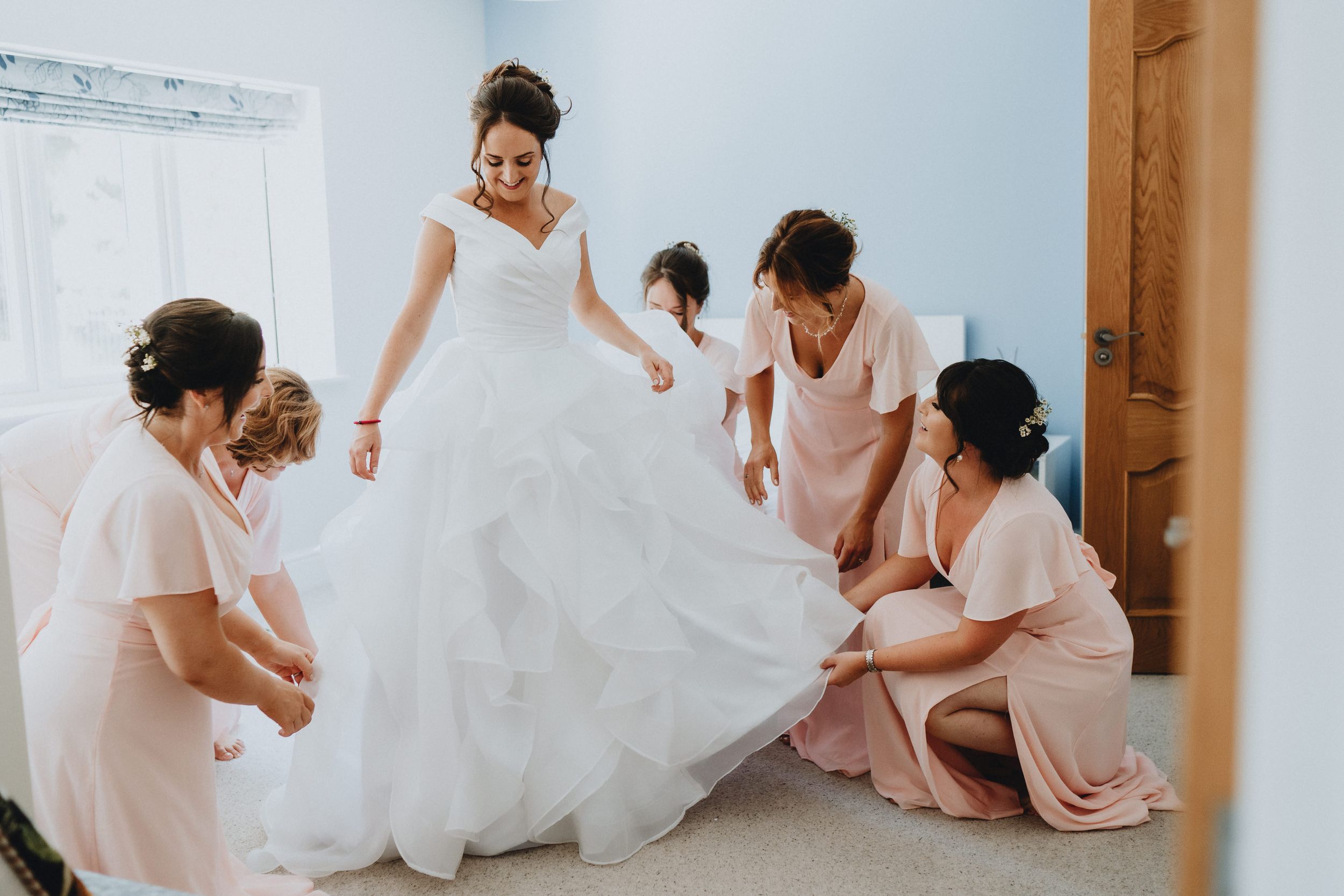 Wedding party in blush pink dresses helps adjust the flowing white ballgown skirt of a bride before the ceremony.