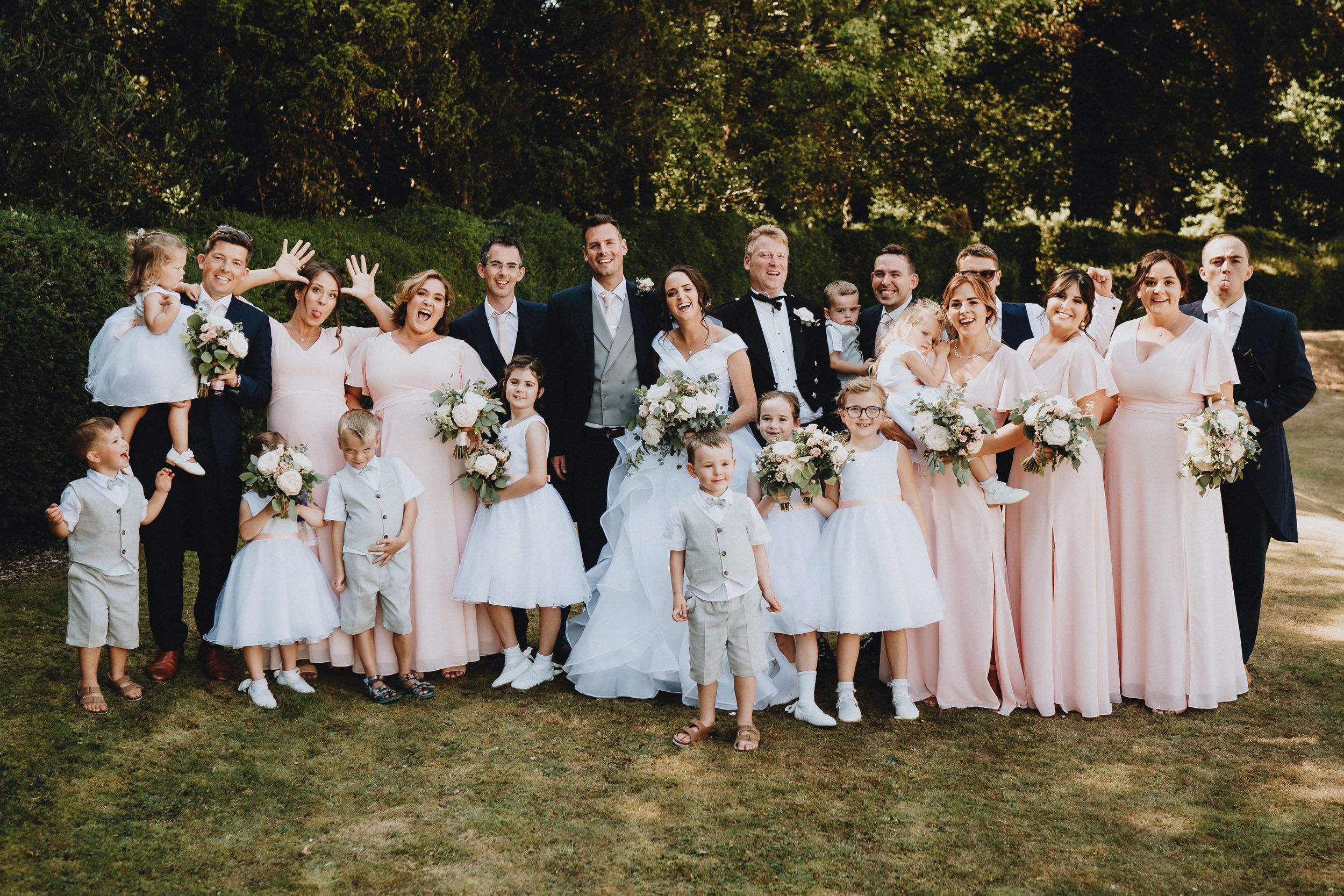 Wedding party gathered outdoors on sunny day wearing pink and black formal attire and holding white floral bouquets.