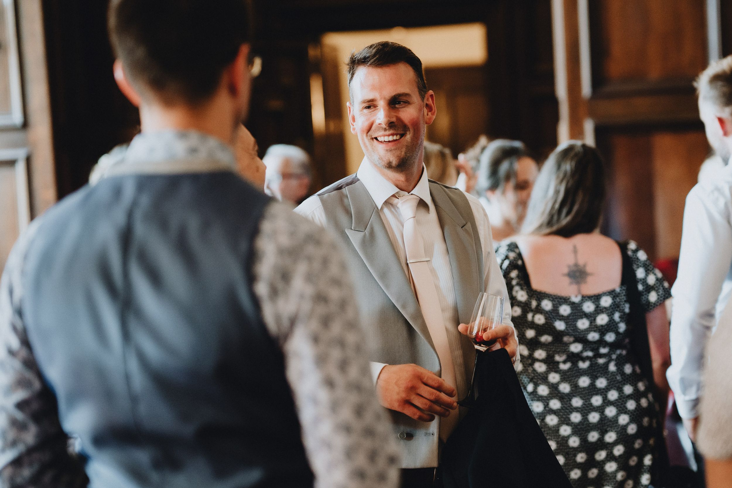 A guest in a light gray suit smiles while mingling at an indoor social gathering.