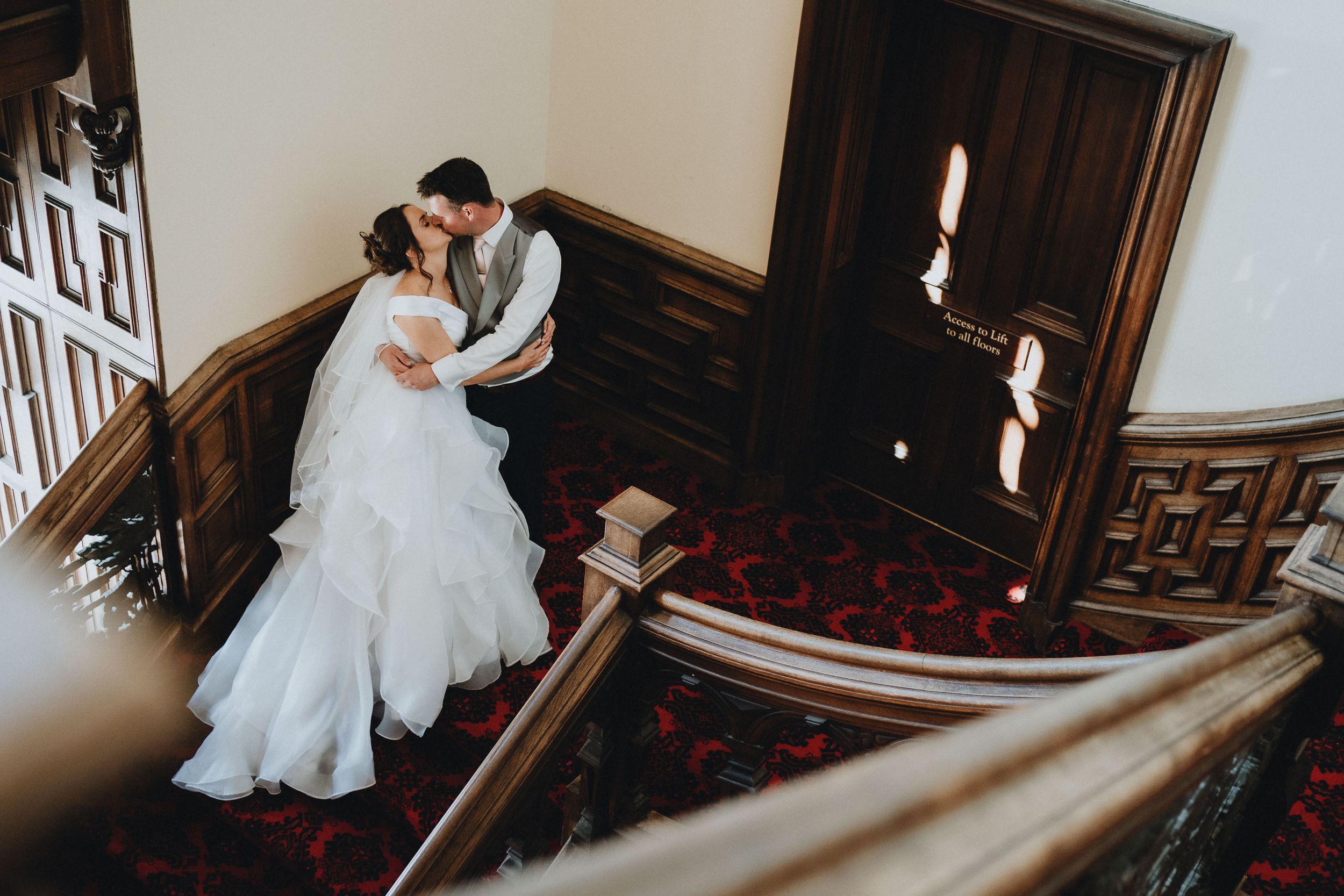 A couple in wedding attire embrace on a vintage staircase at Stoke Rochford Hall with dark wood railings and patterned red carpet.